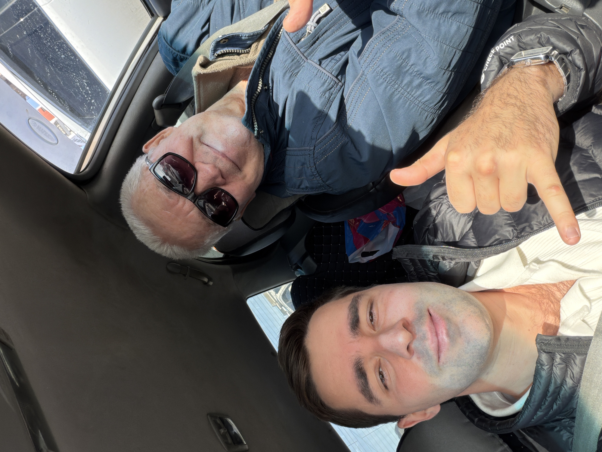 An image, rotated 90 degrees counter-clockwise, depicts two men inside a vehicle on a sunny day. The older man, positioned in the upper part of the frame, has white/grey hair, wears dark sunglasses, and a blue jacket over a beige inner layer. He is smiling faintly. The younger man, in the lower part, has dark hair, a light stubble, and is wearing a black puffer jacket over a white ribbed top. He looks towards the camera with a slight smile and makes a "rock on" or "shaka" hand gesture with his right hand. A silver watch with a square face is visible on his left wrist.
The car's interior, including a light-colored headliner and a seatbelt, is visible. A sunroof or window reflects bright sunlight and shows some minor smudges on the glass. A faint, partial reflection of text, possibly "HAYLO", is visible through this window. On the sleeve of the younger man's black jacket, near his wrist, the white text "CHECK POINT" is visible. A colorful bag is partially seen in the background between the two men. Both individuals appear relaxed and in good spirits, likely taking a selfie or being photographed. The setting is a car interior in Krasnoyarsk, Russia.