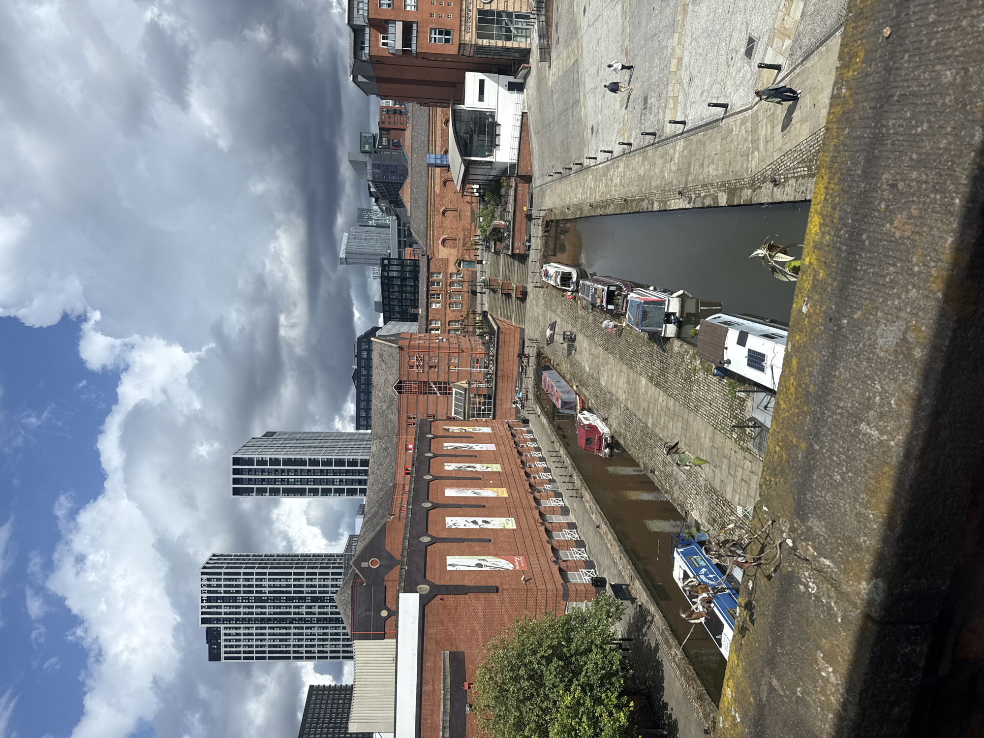 This is a medium-angle, outdoor, eye-level shot of a canal and surrounding urban architecture in Manchester, United Kingdom. The scene is bathed in daylight under a partly cloudy sky. In the foreground, a weathered stone ledge with moss and lichen runs across the bottom of the frame. The canal, a body of dark, still water, occupies the middle ground, lined with moored narrowboats and barges. The boats vary in color and design, with some featuring white hulls and others darker tones, some displaying solar panels. A paved walkway with bollards runs alongside the canal.
Beyond the canal, several red brick buildings with dark tiled roofs and white-framed windows are visible. These buildings have a historical industrial aesthetic. Some have banners with illustrations hanging from their facades. Further in the background, modern high-rise buildings with glass and metal facades dominate the skyline, including two prominent black and white striped towers labeled "UNION".
Several individuals are present in the scene. Two people are walking on the paved area to the right of the canal, a man and a woman. Another person is walking along the canal path on the right. Two individuals appear to be standing near the canal edge, and others are potentially within the buildings or on balconies, but are not clearly discernible. The overall impression is a blend of historical and contemporary urban development along a revitalized waterway.