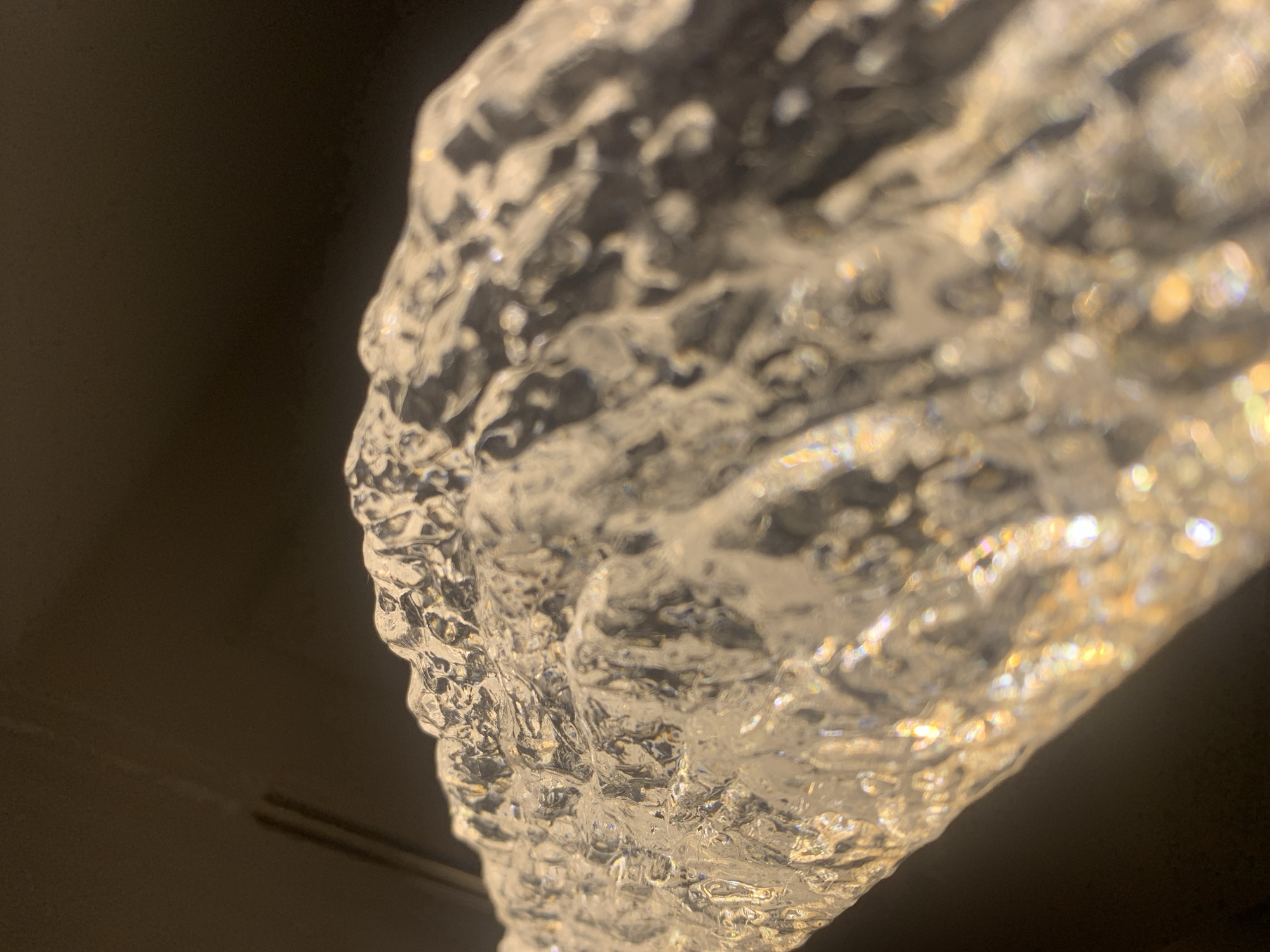 The image is a close-up, abstract shot of a textured, translucent object, possibly made of glass or ice, against a dark background. The object has a rough, uneven surface with many facets that refract light, creating shimmering highlights and a bokeh effect. The dominant colors are clear, white, and golden hues from the reflected light. The background is a deep, out-of-focus black, suggesting a dimly lit environment. There are no visible people, actions, or specific environmental details that would allow for the determination of a location in Vladivostok, Russia. The focus is entirely on the textural and light-play qualities of the object itself.