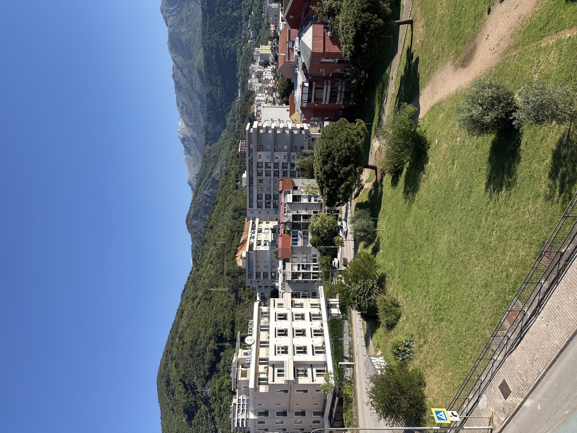 The image shows a cityscape of Unknown, Unknown, featuring a combination of natural and urban elements. The view is from an elevated position, looking down on buildings and a grassy area. A mountain range forms the backdrop to the city. Buildings in the foreground are modern, multi-story structures with white facades and numerous windows. Between the buildings and the road, there is a strip of grass with some trees and bushes. A paved road runs along the bottom right of the image with a metal railing and a street sign.