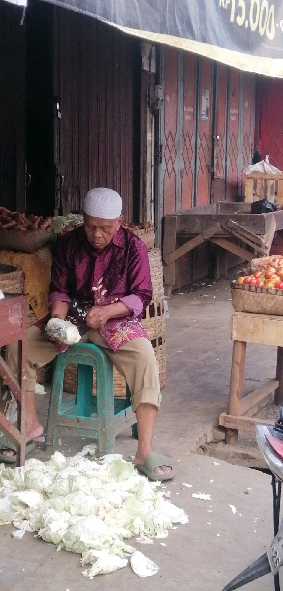 An elderly man wearing a white skullcap and a purple patterned shirt is sitting on a green plastic stool in what appears to be an outdoor market in Ajibarang, Indonesia. He is holding a white object, possibly a large piece of ginger or a root vegetable, and appears to be peeling or preparing it with his hands. In front of him on the ground is a pile of discarded cabbage leaves. To his left is a basket filled with carrots. To his right, on a wooden stand, is a basket filled with tomatoes. Behind him, a large, corrugated metal shutter door is visible, suggesting a shopfront or stall. The overall environment appears to be a bustling marketplace.