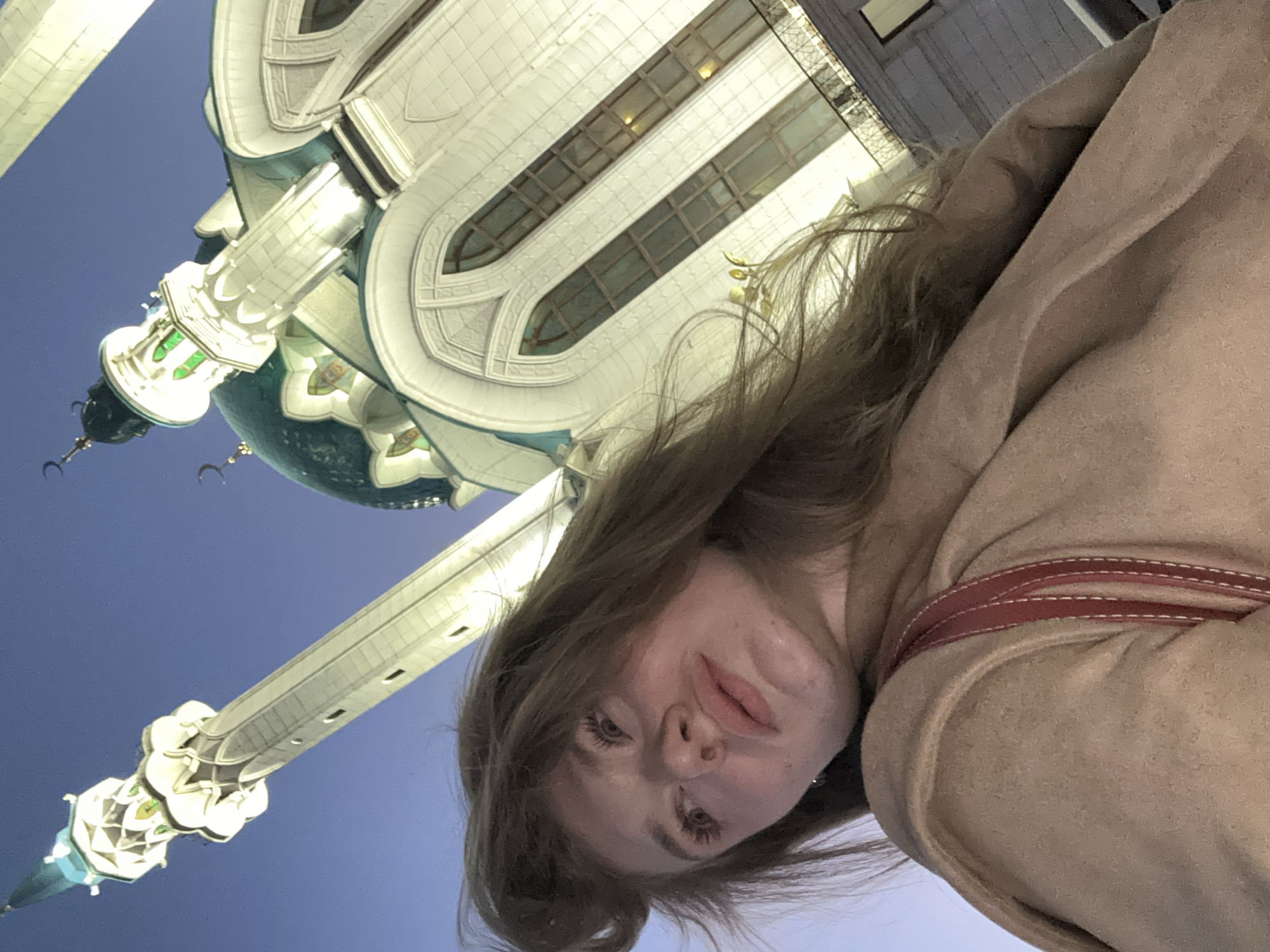 A woman in a tan coat is posing for a selfie in front of the Kul Sharif Mosque in Kazan, Russia. The mosque's minaret and part of the main building are visible in the background. The sky is a gradient of blue.