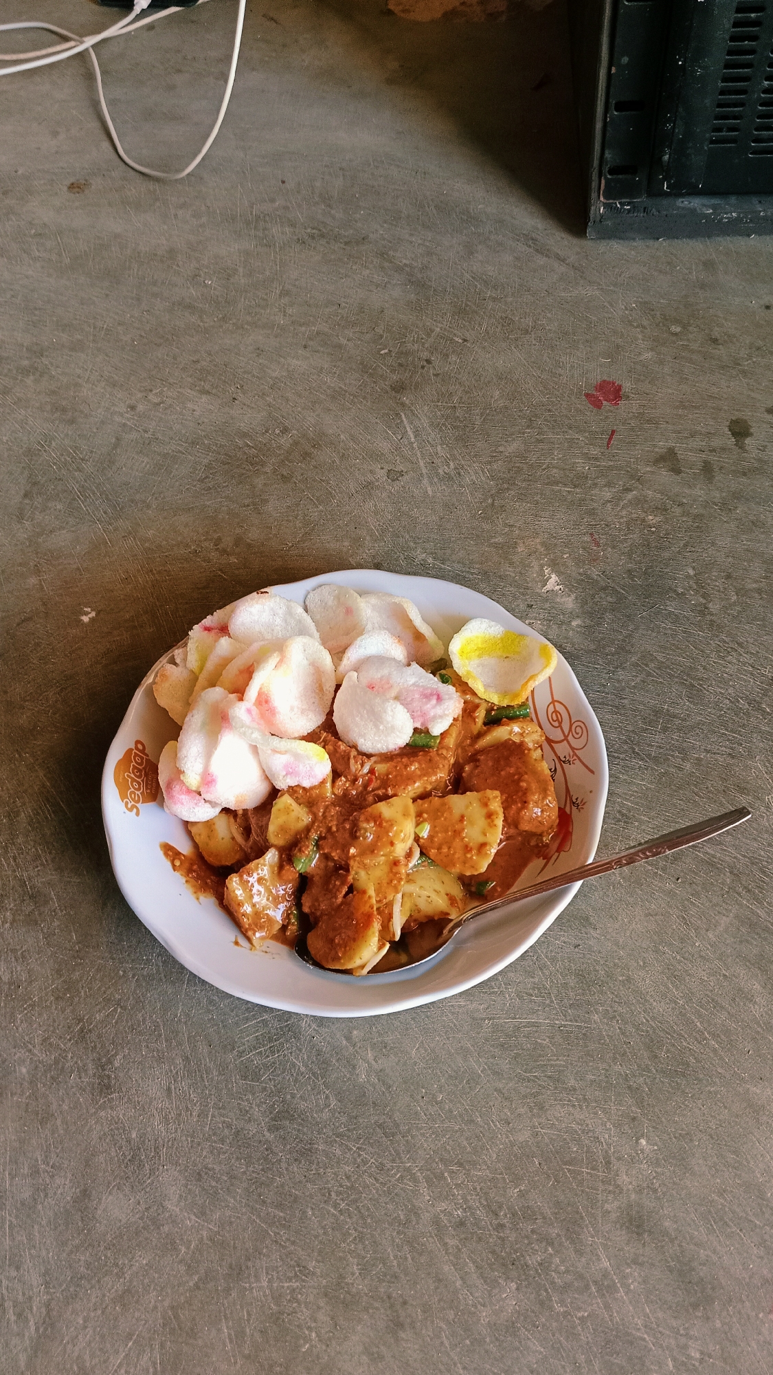 A plate of Indonesian gado-gado, a salad of mixed vegetables, hard-boiled eggs, fried tofu and tempeh, and lontong rice cake, is served with peanut sauce and crackers. The dish is presented on a white plate with an orange and brown patterned rim. A silver spoon rests on the right side of the plate. The plate is positioned on a textured, grey concrete surface that appears to be an outdoor ground. In the background, a white electrical cord snakes across the ground and a black, rectangular electronic device is partially visible. The scene is captured from a slightly elevated, overhead angle. There are no people visible in the image.