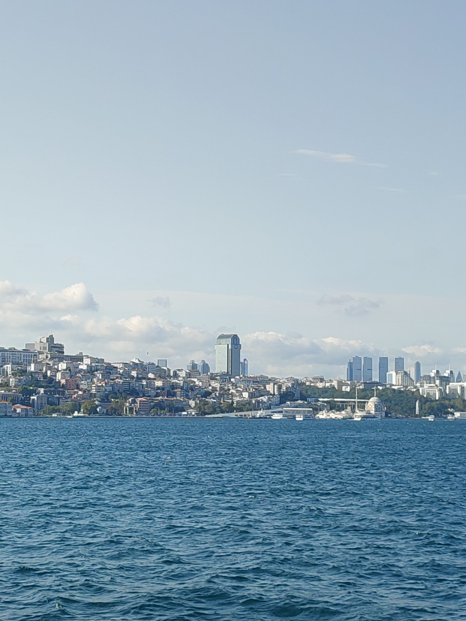 A wide-angle, low-angle, natural light, outdoor, daylight, medium shot captures a vast expanse of deep blue, choppy water in the foreground. A dense urban landscape with numerous buildings, ranging from low-rise to high-rise structures, fills the midground. Among the buildings, a distinctive tall, rectangular skyscraper with a green, reflective facade stands out, as do three identical, closely grouped, rectangular skyscrapers. A dome-shaped building with a minaret is also visible. The cityscape recedes into the distance, meeting a hazy, pale blue sky dotted with sparse, wispy white clouds. The overall scene depicts a coastal city with a mix of old and modern architecture. Based on the characteristic skyline featuring the distinct green skyscraper and the overall topography, the location is likely the Bosphorus strait in Istanbul, Turkey, looking towards the European side.
