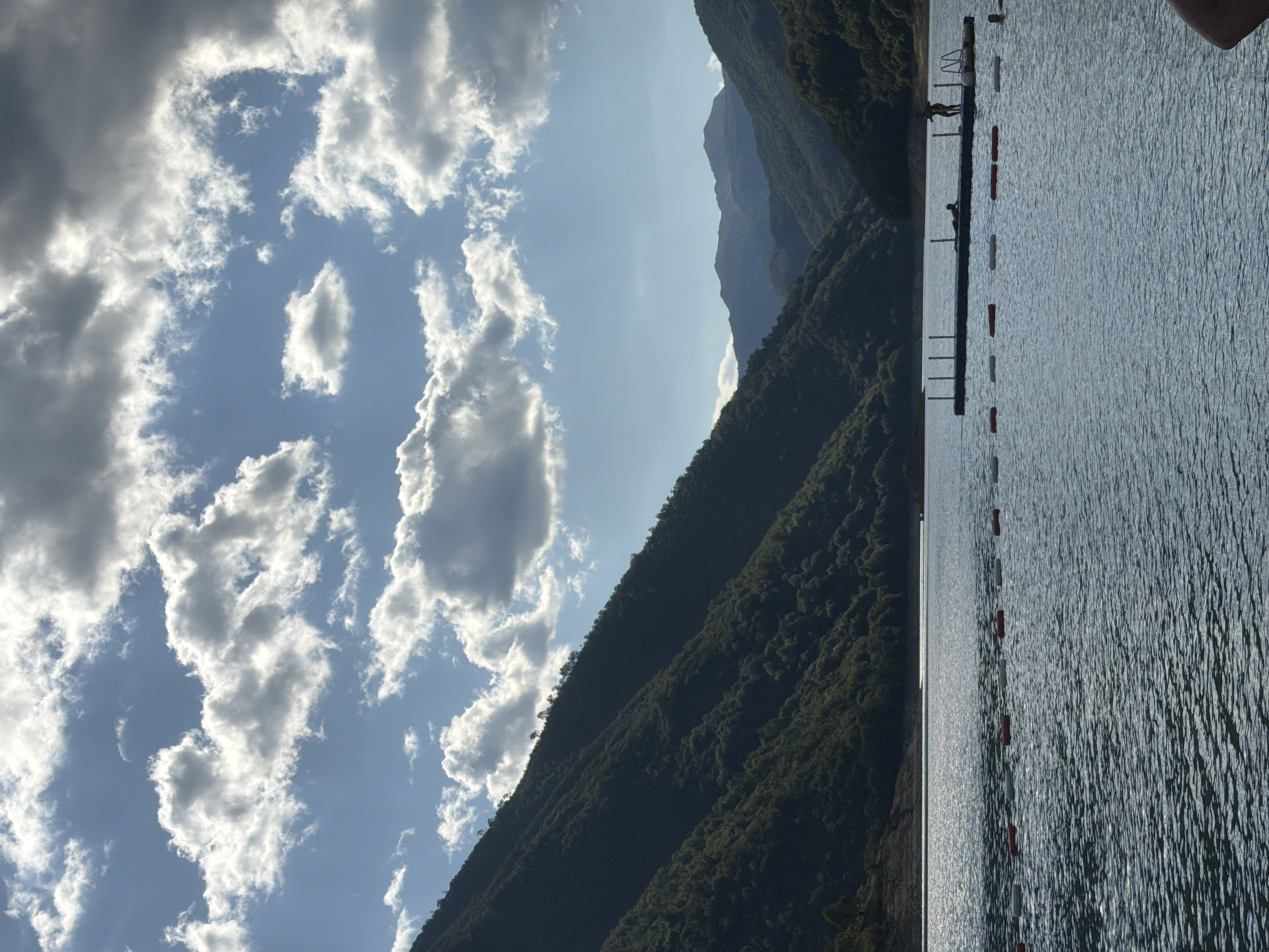 The image depicts a serene lake surrounded by lush green mountains under a dramatic sky filled with white and gray clouds. In the foreground, a calm body of water reflects the sunlight, with a line of buoys marking a section of the lake. In the mid-ground, a floating platform is visible, with two figures standing on it and one lying down. Another person is partially submerged in the water near the platform. The background features the imposing, tree-covered slopes of mountains that flank the lake, with more distant mountain ranges fading into the haze. The overall scene suggests a recreational area, likely a popular spot for swimming and enjoying nature. The location is the Piva Lake near Plužine, Montenegro, a man-made reservoir known for its stunning natural beauty and clear waters.