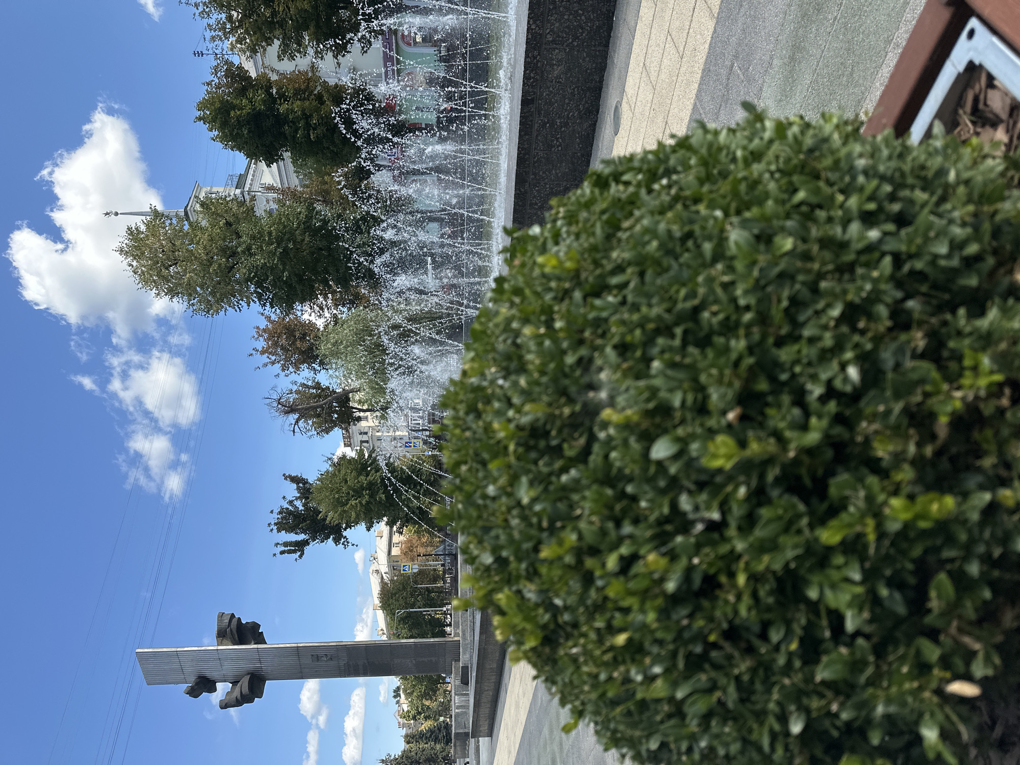The image captures a city square in Raivka, Ukraine on a bright, sunny day with scattered clouds. In the foreground, a meticulously trimmed, spherical green bush occupies the lower left portion of the frame. Behind the bush, a long, linear fountain sprays water upwards, creating a shimmering curtain. To the left of the fountain, a tall, modern monument with abstract, angular elements stands prominently. In the background, several trees with green and some autumnal foliage line the square, interspersed with buildings. One building, located to the right of the monument and behind the fountain, features a distinctive spire topped with a star. Power lines are visible stretching across the blue sky. The ground of the square appears to be paved with light-colored stones. There are no people visible in the image.