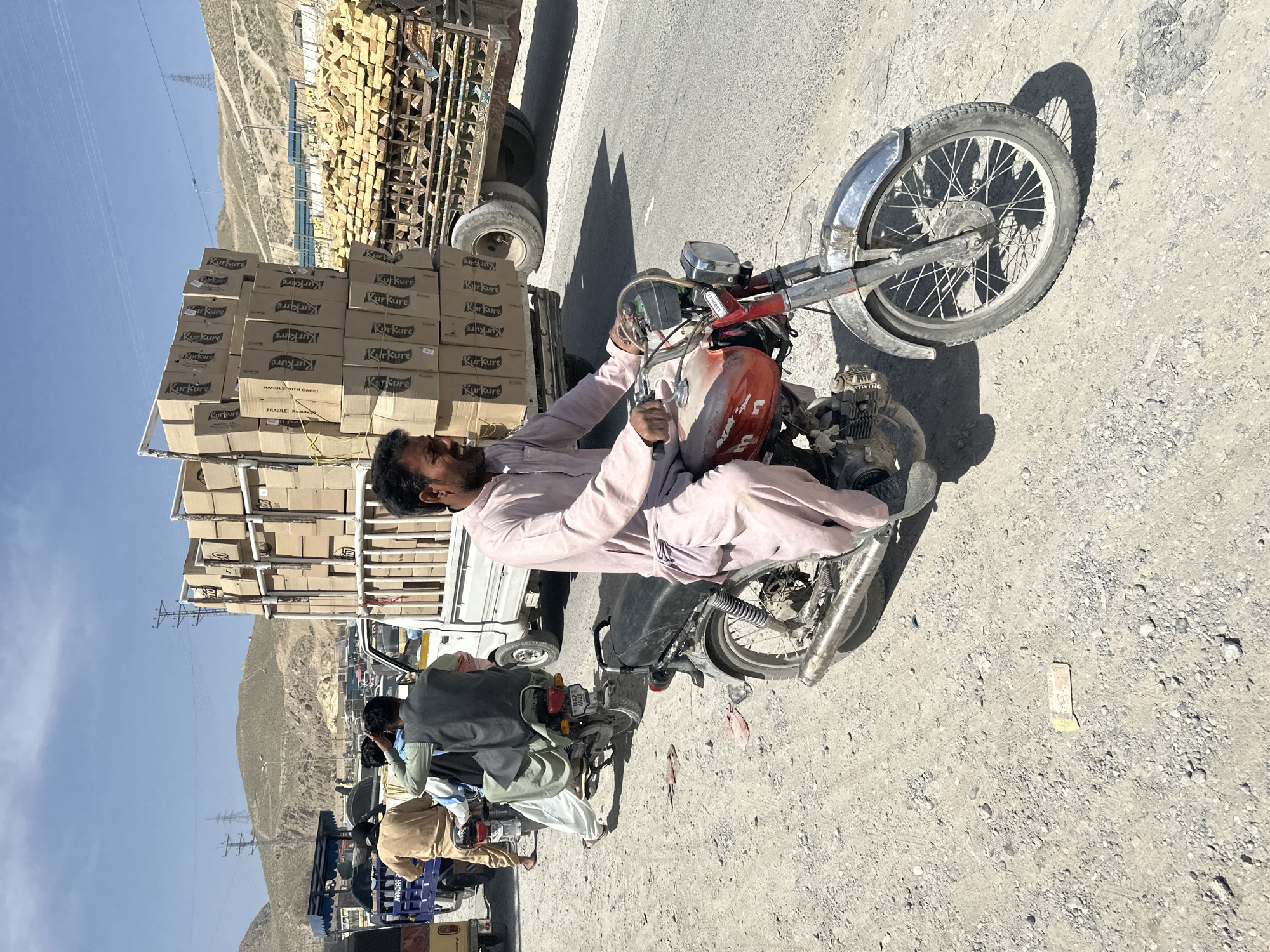 The image captures a sunny, dusty roadside scene, likely in a mountainous, arid region. A man, dressed in a light-colored shalwar kameez, sits astride a red and silver motorcycle in the foreground. He holds the handlebars, his expression neutral, possibly indicating waiting or resting.
Adjacent to the man's motorcycle is a white utility truck, heavily loaded with numerous cardboard boxes stacked high. Most of these boxes are clearly branded "Kurkure," and some feature text such as "HANDLE WITH CARE!" and "FRAGILE!". The truck appears to be either parked or moving slowly.
In the background, the road continues, revealing other people and vehicles, including additional motorcycles and what appears to be another truck. One visible license plate reads "MAP 4028," and "JAANDAR" is visible on another vehicle. The landscape consists of barren, light-colored mountains under a clear blue sky, with power lines and utility poles stretching across the terrain. Strong, sharp shadows cast by the subjects and vehicles suggest bright midday sunlight. The scene depicts activity along a transportation route, possibly a delivery or a stop in a remote area.