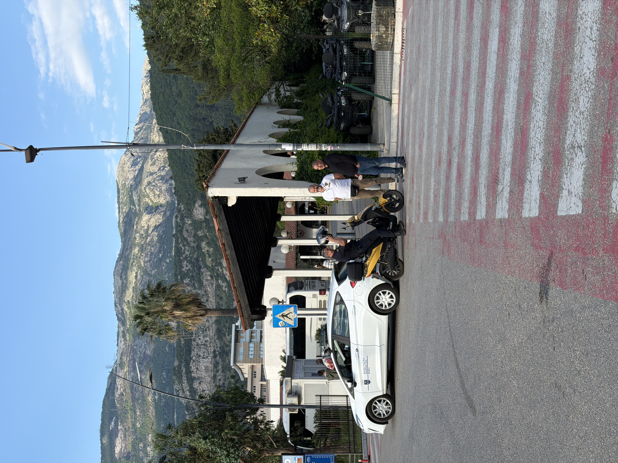 The image shows a street scene in Budva, Montenegro. A white car with official markings is parked on the side of the road. A man in uniform is sitting on a yellow scooter next to the car. Three men are standing on a pedestrian crossing nearby. A building with white arches and lush greenery in the background is visible. The street has a view of mountains and a blue sky with scattered clouds. Several ATVs are parked in the background.