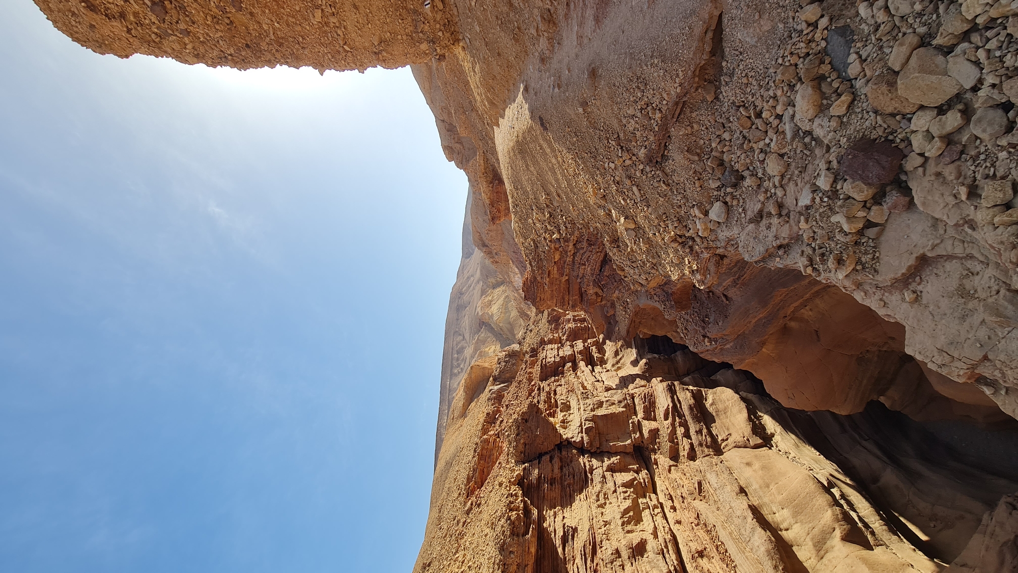 The image, taken in Hevel Eilot Regional Council, Israel, presents a low-angle perspective of a canyon or ravine. The foreground features rugged, textured rock formations in shades of brown and orange. To the right, the canyon walls appear close, with visible sedimentary layering and small caves or overhangs. Above and to the left, a patch of bright blue sky is visible, suggesting a sunny day. The top of the frame shows more rocky overhangs, casting shadows on the canyon walls. The scene is devoid of people or animals, focusing solely on the geological features.