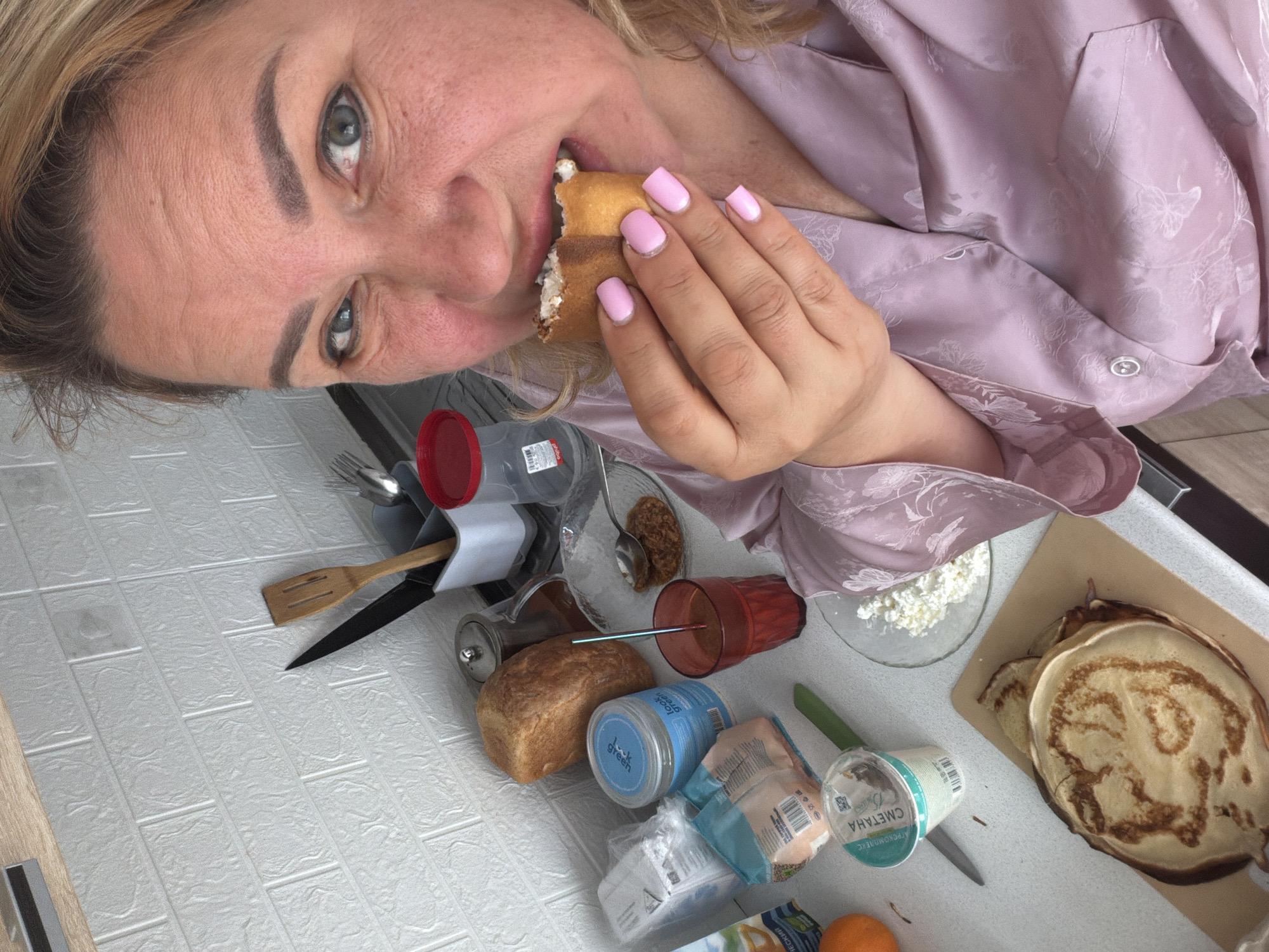 A woman in a light purple pajama-like shirt is eating a sandwich with a white filling. Her nails are painted a light pink color. The scene takes place on a white tile countertop. There is a loaf of bread, a can of "Luck Green" product, a glass bowl with brown substance inside, a red glass cup, a bowl of cottage cheese, a container of sour cream labeled "СМЕТАНА", a plate of pancakes, silverware, a wooden spatula, a knife, an orange, and packaged items. Based on the label "СМЕТАНА" and the Cyrillic writing on other packages, the location is likely in a Russian-speaking region. Given the label "СМЕТАНА" (sour cream) and that is written in Russian, it is likely to be somewhere in Russia, Ukraine, or Belarus. It is not possible to confirm the location in Leningradskaya, Russia, with the information present in the image.