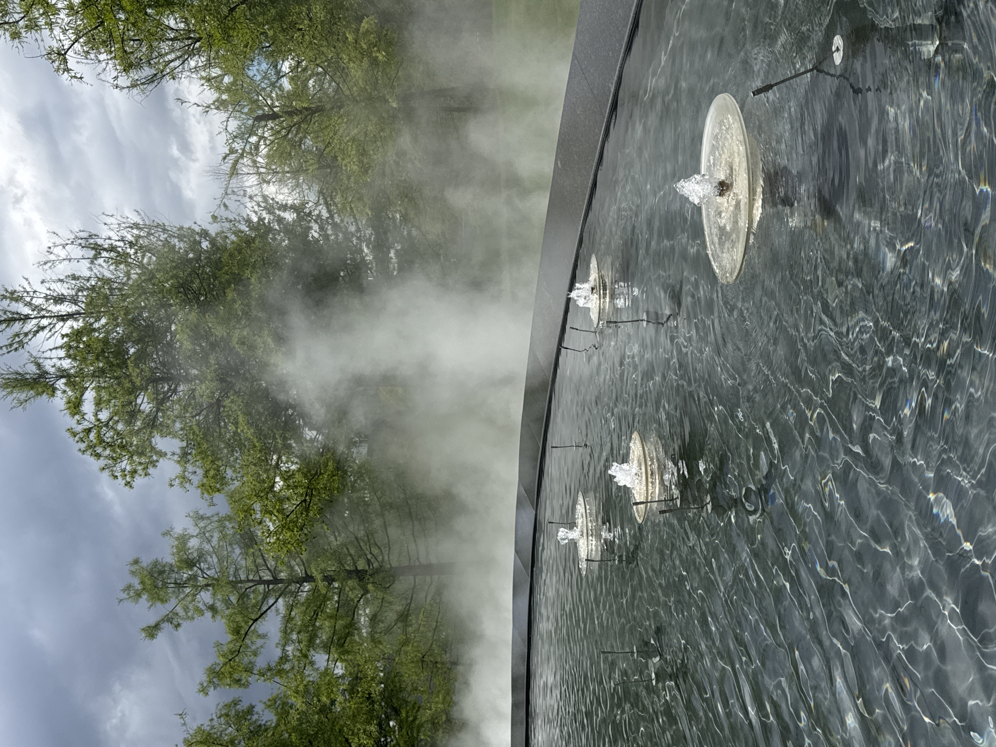 The image shows a fountain with water jets spraying upwards. The fountain is situated beside a park-like area with trees and mist. The water in the fountain has reflective ripples. The location is Krasnodar, Russia.