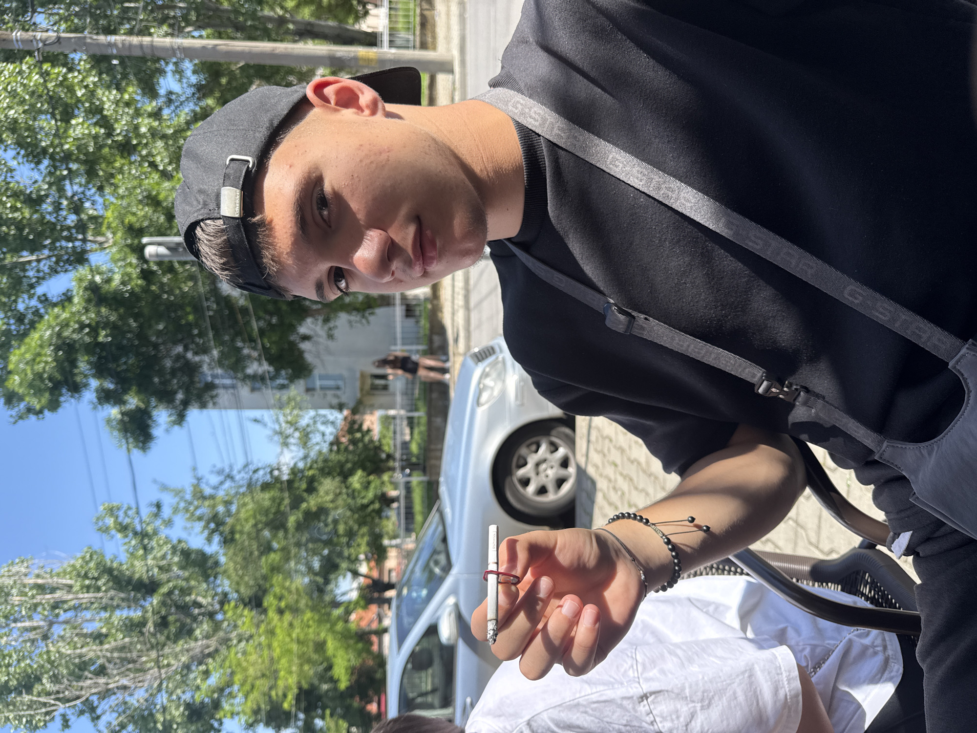 A young man is holding a cigarette with a red rubber band around it in his right hand. He is wearing a black baseball cap and a black t-shirt with a gray strap across his chest. He is sitting at an outdoor table. A silver car is parked behind him. The location is Elhovo, Bulgaria. Trees and buildings are visible in the background.