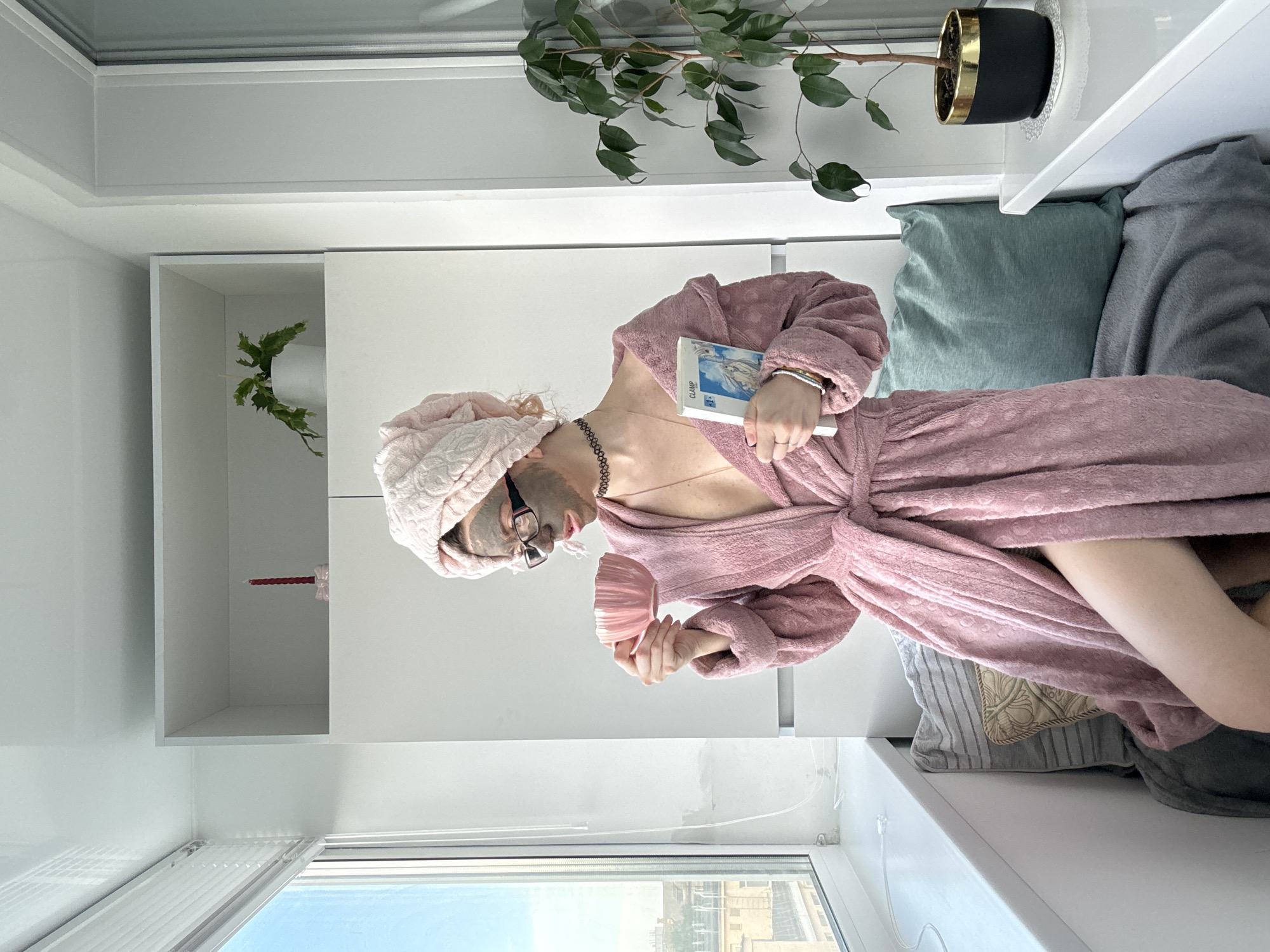 In the city of Unknown, Unknown, a young woman with a pink towel wrapped around her head and wearing a pink terrycloth robe with a clay mask on her face sits by a window. She holds a pink mug in her right hand and a book in her left. She is wearing glasses and a black choker necklace. Beside her, on a windowsill, is a potted plant. The room appears to be an interior space with built-in shelving and a window with blinds.