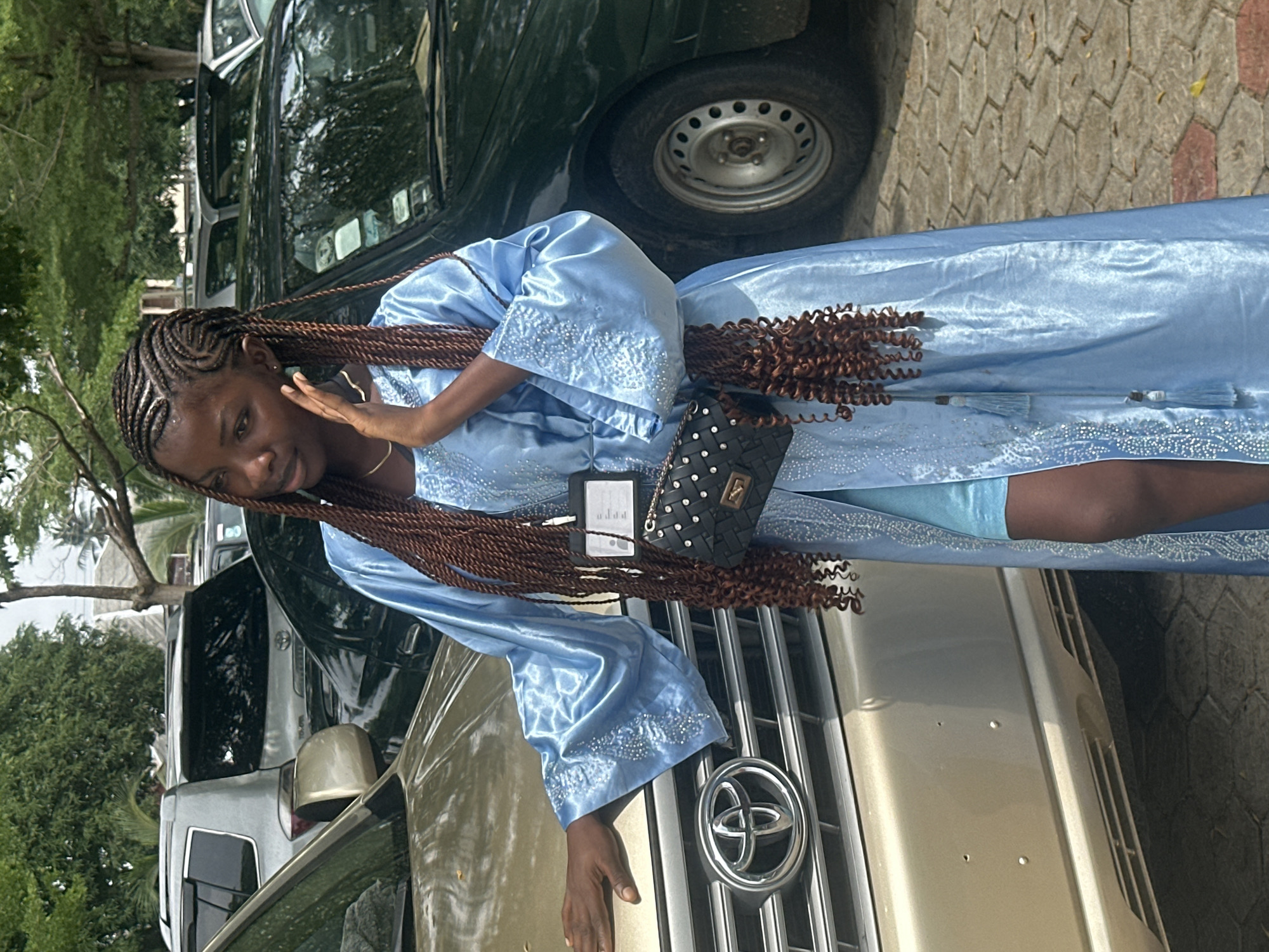 A young Black woman with braided hair stands in front of a gold Toyota SUV. She is wearing a light blue, shimmering robe with intricate beading on the sleeves and hem. A black quilted purse with silver studs hangs from her shoulder. She has her right hand resting on the car's hood and her left hand on her cheek. The scene appears to be in a parking lot, with other vehicles visible in the background, including a dark green car and a silver SUV. The ground is paved with interlocking bricks. The environment suggests a warm, possibly tropical climate with lush trees and foliage. The image was taken on a bright, overcast day. The location is Itoga, Nigeria.
