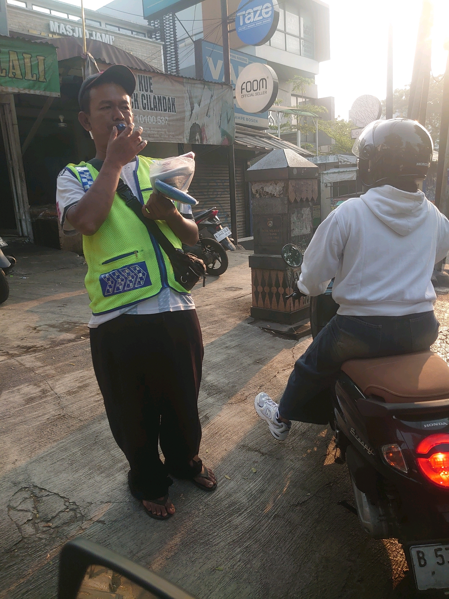 In South Jakarta, Indonesia, a man wearing a black cap, a white t-shirt, and a neon green vest with reflective trim stands on a concrete sidewalk. He is holding a plastic bag containing a blue object and has a blue item near his mouth. To his right, another person wearing a white hoodie and dark jeans sits on a dark-colored scooter. The scooter's taillight is illuminated. The background shows a building with signage, including "MASJID JAMI" and "TAZE." The sun is low in the sky, casting long shadows. The scene appears to be a street or marketplace.