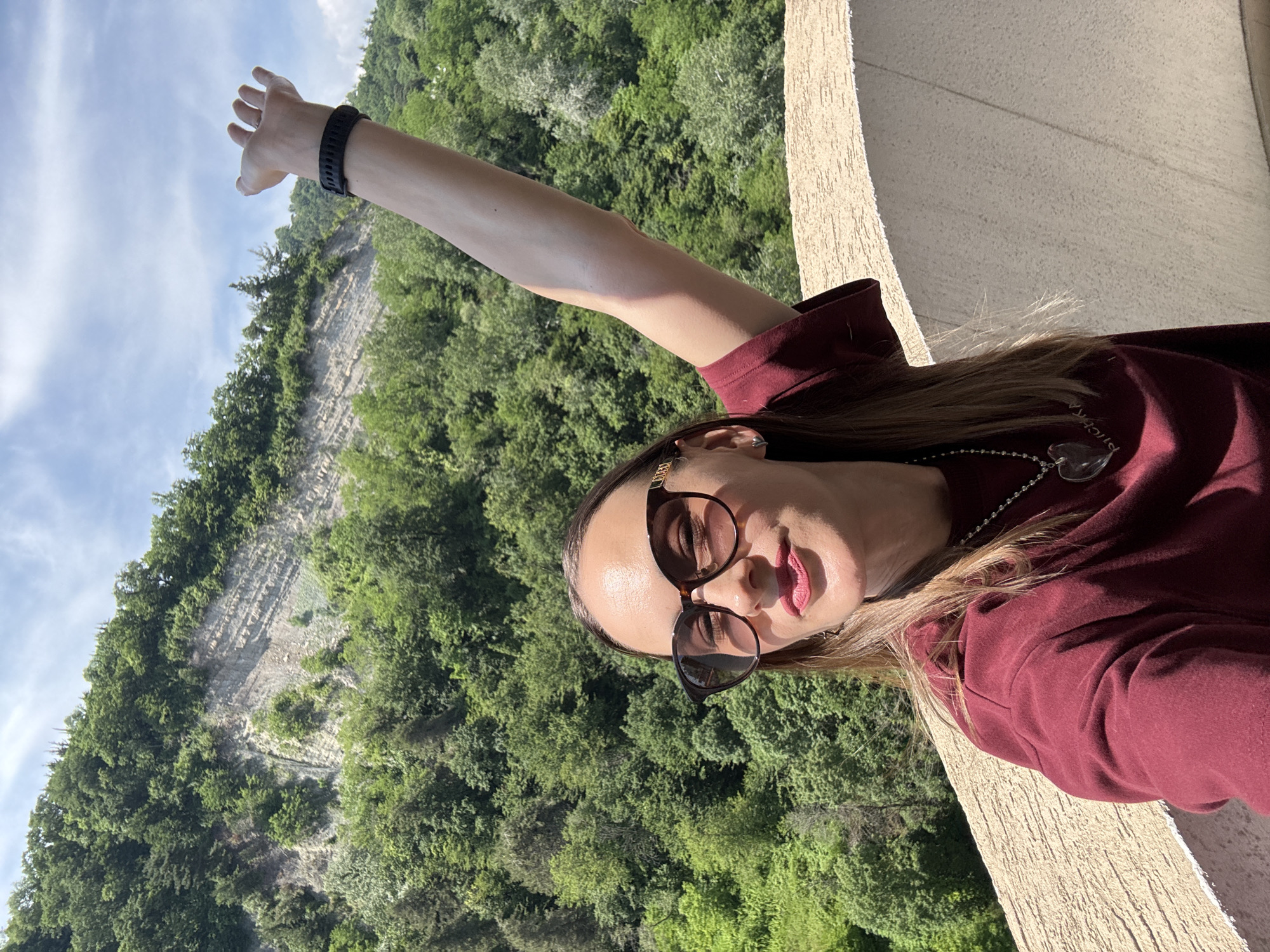In Sochi, Russia, a young woman stands on a balcony with a lush, green hillside and a rocky cliff face in the background. She is wearing a maroon t-shirt, large brown-rimmed sunglasses, and a silver necklace with a heart-shaped pendant. Her right arm is raised, with her hand open. The sky above is blue with scattered white clouds. The balcony railing is a light, textured beige.