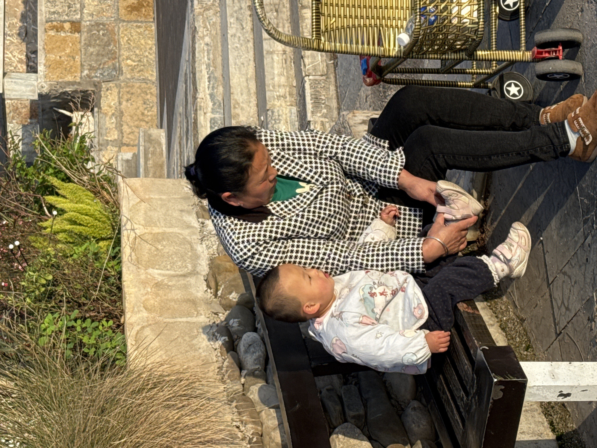 A woman in a black and white checkered jacket and black pants sits on a black wooden bench in Zhuoshui, China. She is putting a white and pink shoe on a child in a white patterned onesie and dark pants who is lying across the bench. The woman wears brown shoes and a silver bracelet on her left wrist. A golden stroller sits on the stone pathway behind the woman. To the left of the bench is an area with grass and rocks. The bench appears to be near a set of stone stairs. The day appears to be sunny.