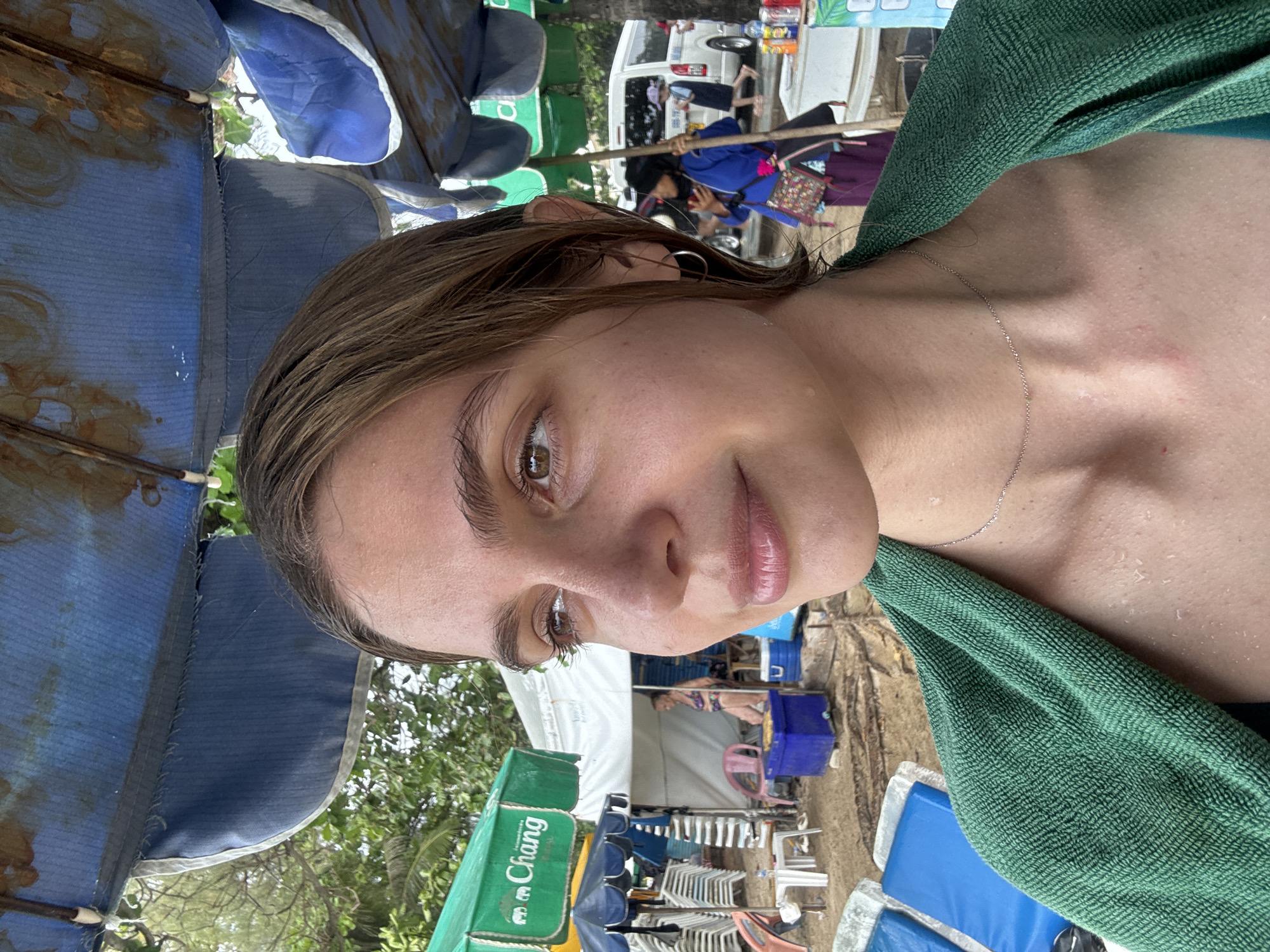 A close-up photograph taken at what appears to be a beach, featuring a woman with brown hair and eyes, wearing a green towel draped around her neck. The background suggests an outdoor location, likely a beach in Patong, Thailand, evidenced by the presence of beach umbrellas, lounge chairs, and commercial branding (Chang beer). A white van is visible in the distant background. The woman is looking at the camera.