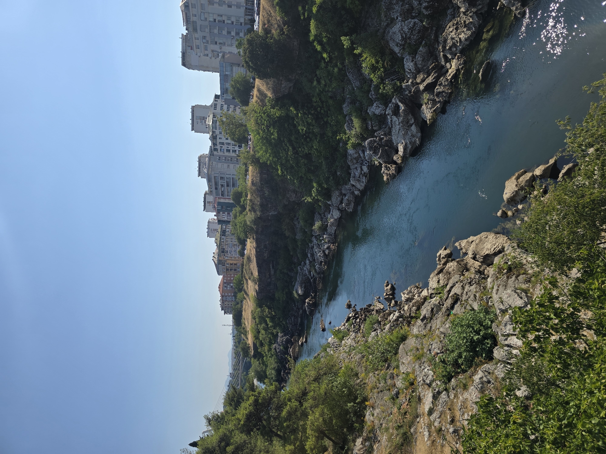 A clear, sunny day is depicted with a deep blue sky above. A wide, calm river flows through a rocky gorge, with dense green foliage clinging to the rocky banks on the left side of the frame. The river's surface is disturbed by ripples and reflects the sunlight in shimmering patches. On the right bank, the gorge continues, lined with rocks and trees. Beyond the gorge, a city skyline rises, featuring several multi-story buildings. A bridge spans the river in the distance, connecting the city. The overall scene is a juxtaposition of natural beauty and urban development.