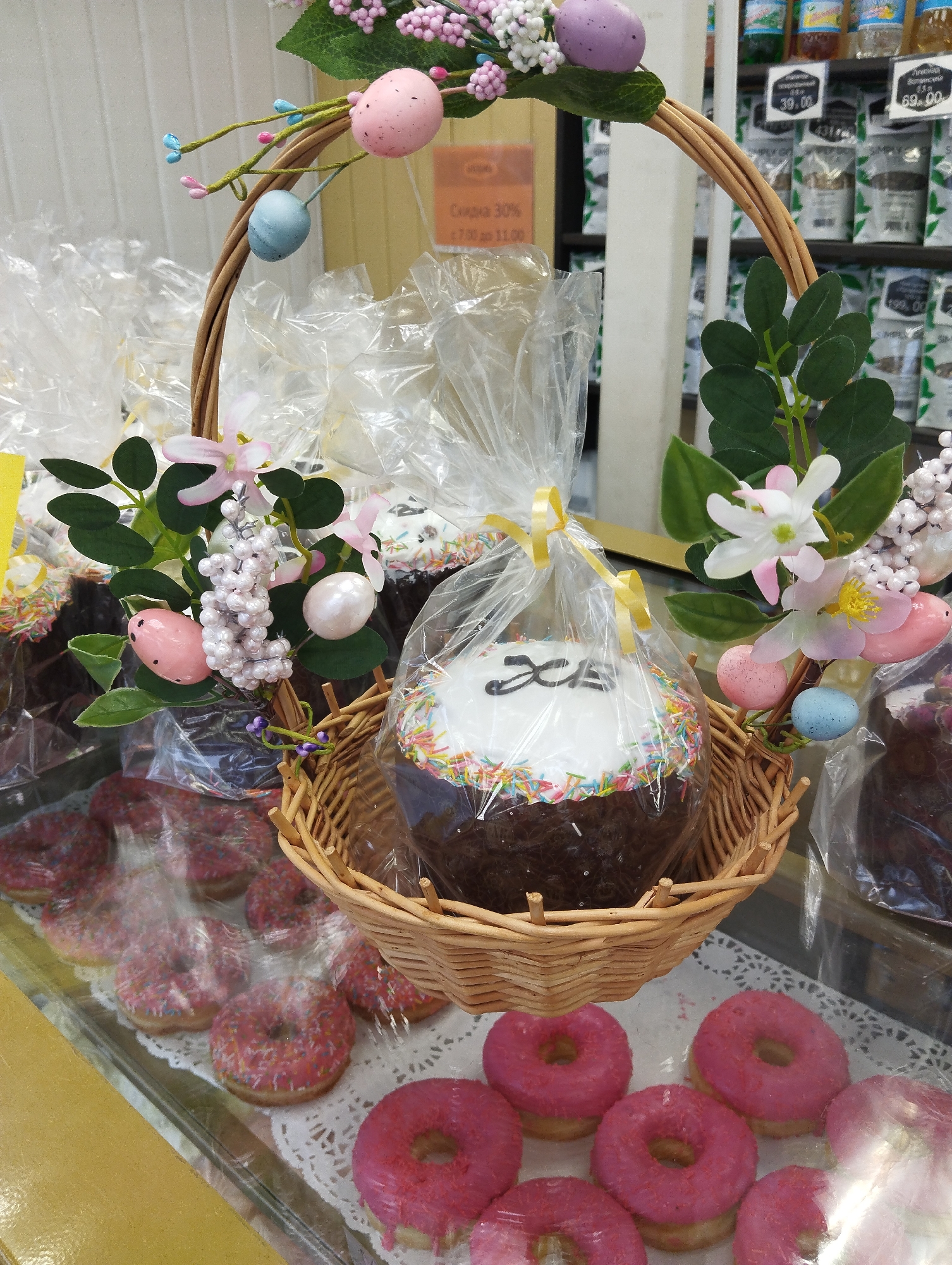 The image shows a retail display in Votkinsk, Russia, featuring baked goods for sale. A wicker basket, decorated with artificial flowers and pastel-colored eggs, holds packaged Easter cakes. The cakes are topped with white icing and colorful sprinkles. Below the basket, glazed pink donuts are arranged on a doily. Behind the basket, shelves are stocked with packaged goods and price tags are visible.