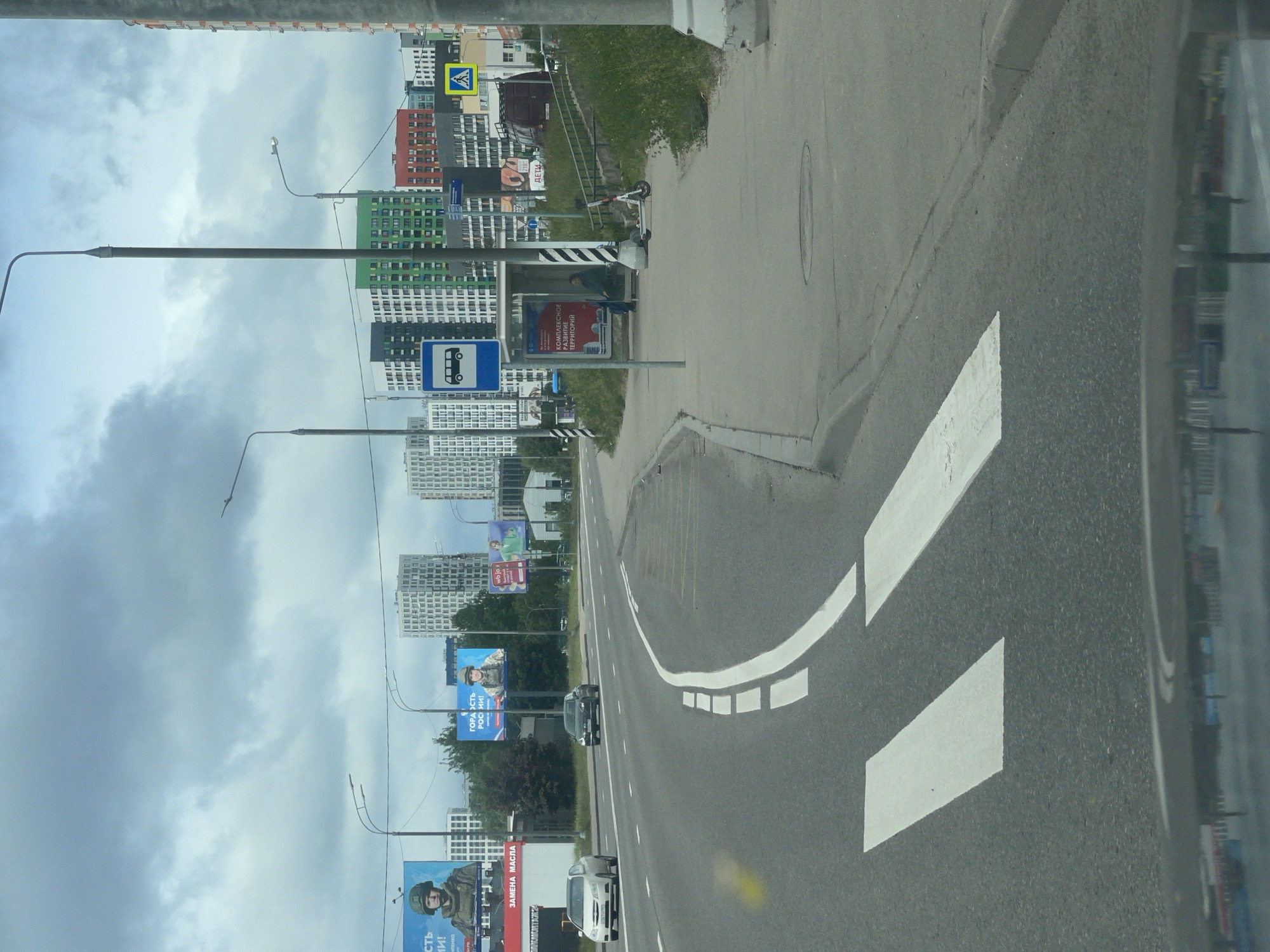 The image shows a street scene in Moscow, Russia, on a cloudy day. The road has white lane markings and arrows indicating direction. Several cars are visible on the road. Alongside the road is a sidewalk with a bus stop shelter. A person is seated at the bus stop. A row of modern apartment buildings and various billboards line the street. Lampposts are also visible along the road. Military-themed billboards promoting Russian patriotism can be seen in the foreground.