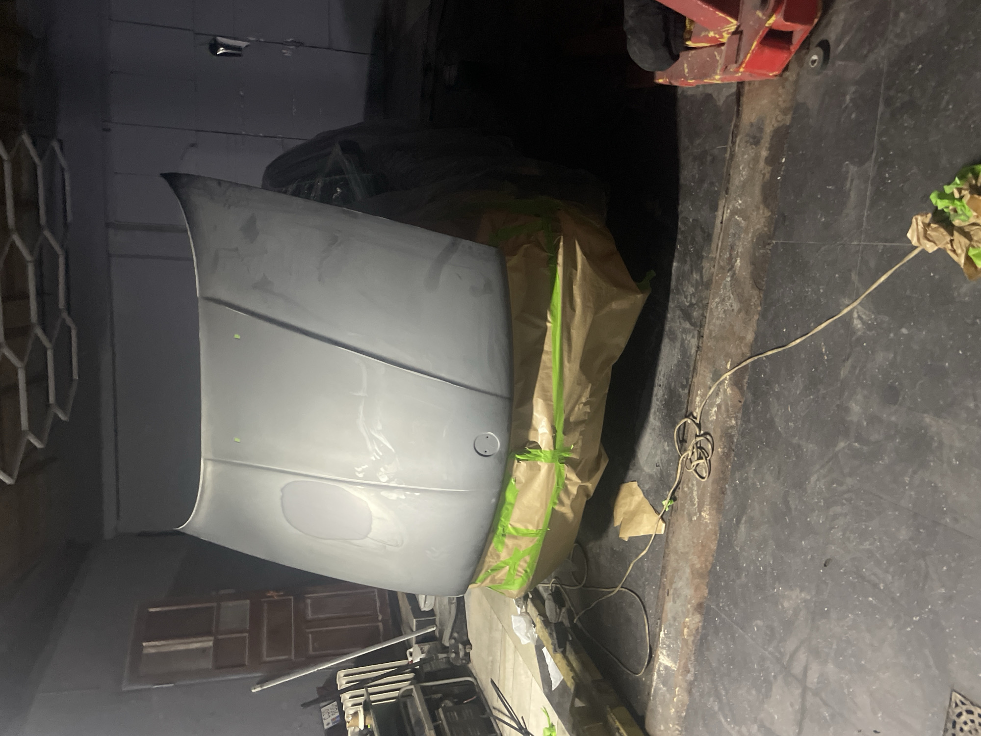 A car hood, covered in primer and partially taped with brown paper and green painter's tape, is propped open in a dimly lit garage. The hood is the primary subject, angled upwards and towards the viewer. Underneath the hood, the engine compartment is shrouded in plastic sheeting. The garage floor is dark and appears to be concrete, with a rusted metal strip running across it. In the background, a wooden door with multiple panels is visible, along with various pieces of equipment and a hexagonal ceiling structure. A red pallet jack is partially visible on the right side of the image. The overall scene suggests a car repair or restoration project in progress.