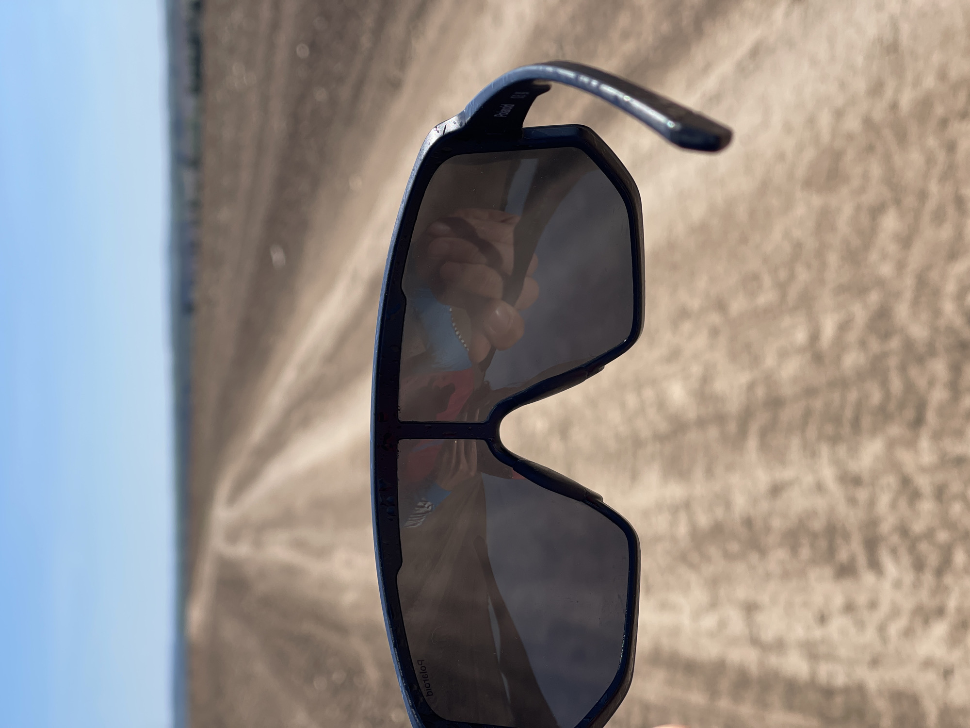 The image depicts a pair of black sunglasses held in front of a blurred background. The glasses are reflecting a person's hand and a hint of clothing. The background is a landscape that transitions from a light blue sky on the left to a tan, seemingly plowed field or terrain, with visual lines converging into the distance. "Polaroid" is visible in reverse on one of the lenses. The location is Perevoznoe, Russia.