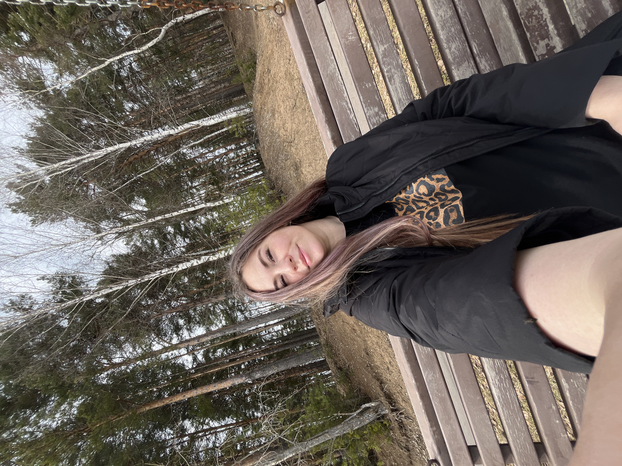 A young woman is lying on a wooden park bench in a park in the city of Chaikovsky, Russia. She is wearing a black jacket over a black leopard-print shirt. The park has a mix of birch and pine trees. A metal chain hangs above the bench. The ground beside the bench is covered in dry grass and leaves.