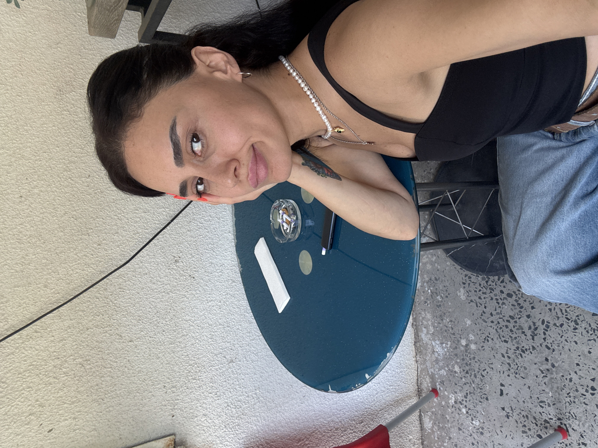 A woman is seated at a small, round, teal table on a concrete patio. She is facing the camera, with her right hand resting on her cheek. The woman has dark hair, dark eyes, and is wearing a black tank top and light blue jeans. Around her neck, she wears a pearl necklace. On the table, there is an ashtray with cigarette butts, a pen, and a white paper. The background consists of a textured white wall and a part of a black metal chair. The floor is speckled gray concrete.