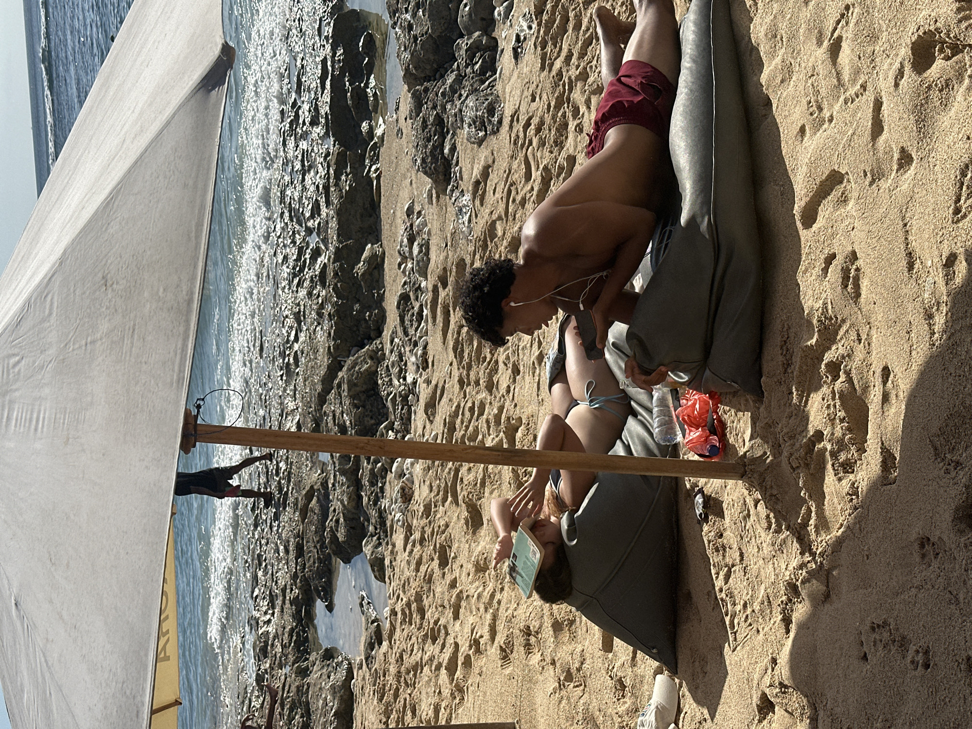 In Kuta Selatan, Indonesia, under the shade of a large white umbrella on a sandy beach, two individuals are relaxing by the ocean. A female is lying on her back on a dark gray beanbag, with a book resting on her face. She is wearing a bikini. Beside her, a male is lying on his stomach on another dark gray beanbag, wearing red swim trunks and white earbuds. He is looking at his phone. The ocean waves are visible in the background, breaking against a rocky shoreline. The lighting suggests it is daytime, with sunlight casting shadows on the sand.