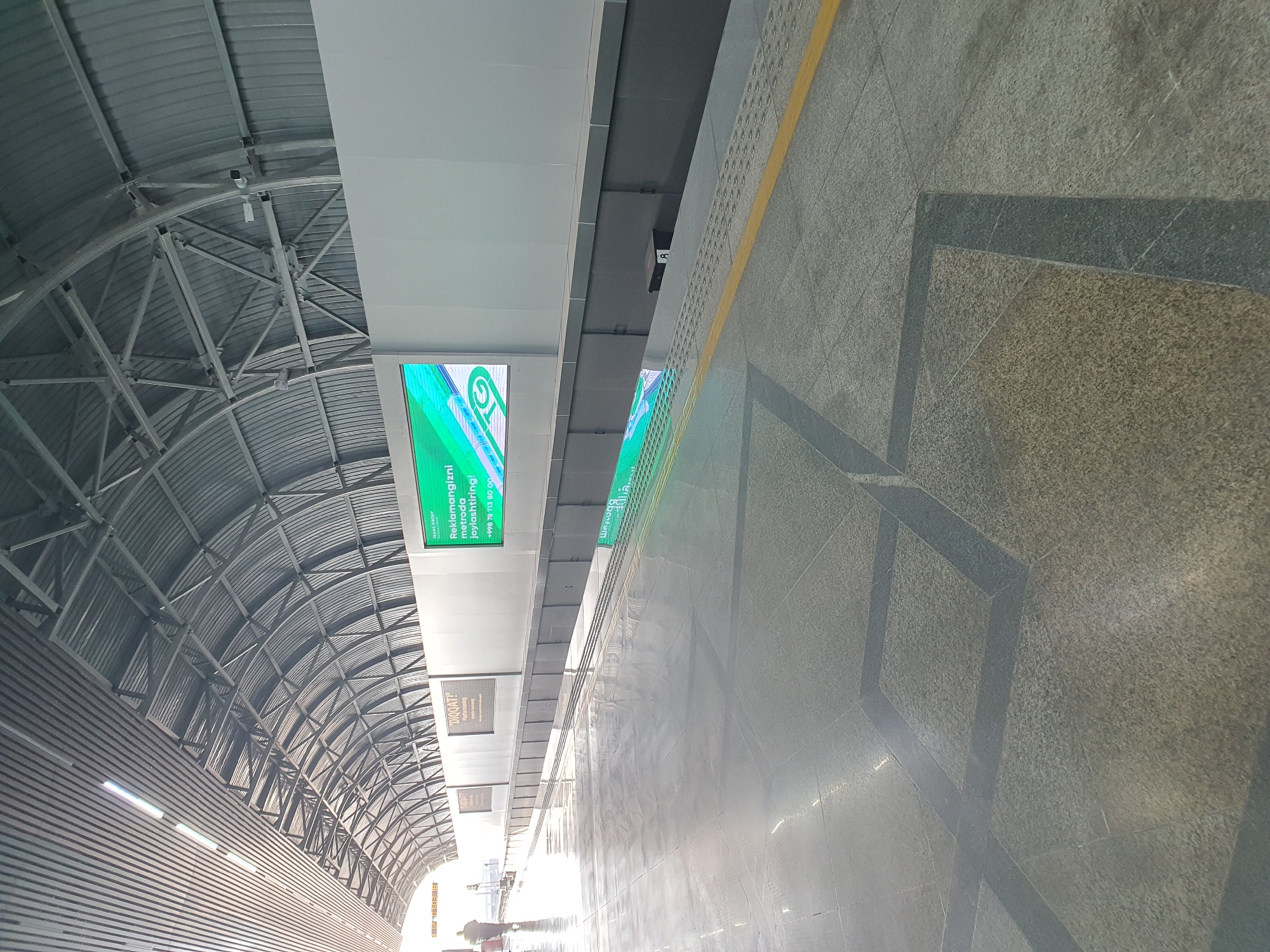 The image depicts the interior of a modern, spacious metro station in Tashkent, Uzbekistan. The station features a high, arched ceiling constructed of metal beams and corrugated metal panels, with fluorescent lighting integrated into the structure. The platform is paved with large, polished gray tiles, interspersed with dark gray geometric patterns. A yellow tactile paving strip runs along the edge of the platform.
On the wall behind the platform, a large digital screen displays a vibrant green advertisement with the text "Reklamangizni metrodan joylashtiring!" and a phone number "+998 78 113 80 00". To the left of this screen, there are two smaller, rectangular signs with "DIQQAT!" written on them, suggesting a warning or important notice.
A lone figure, dressed in dark clothing, is visible walking away from the camera down the platform, adding a sense of scale and emptiness to the scene. The environment is clean and well-lit, with the polished floor reflecting the overhead lighting and the digital display. The overall impression is one of a contemporary and functional public transportation hub.