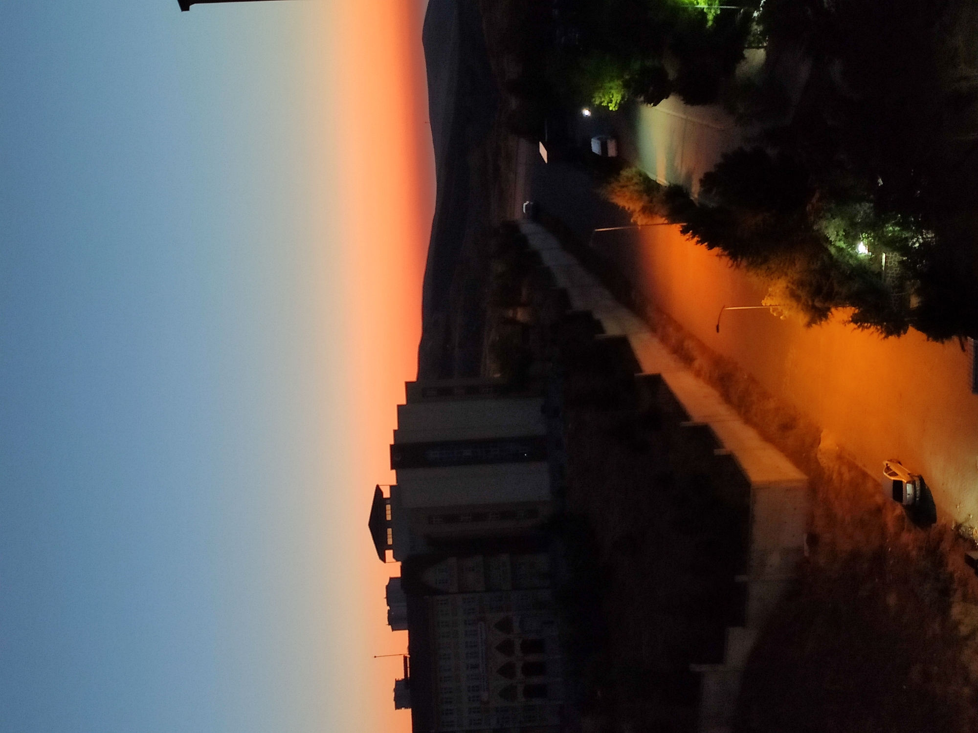 This image captures a dusk scene in the city of Etimesgut, Turkey. In the foreground, a dark, multi-story building with arched windows is visible on the left. A road runs alongside the building, illuminated by a street light, casting an orange glow. Further down the road, two vehicles, a white van and a silver car, are parked. The landscape behind the buildings consists of rolling hills that fade into the twilight sky, which displays a gradient of blue and orange hues. Trees and foliage are scattered throughout the scene, with some illuminated by artificial lights. The overall impression is one of a quiet, urban evening.