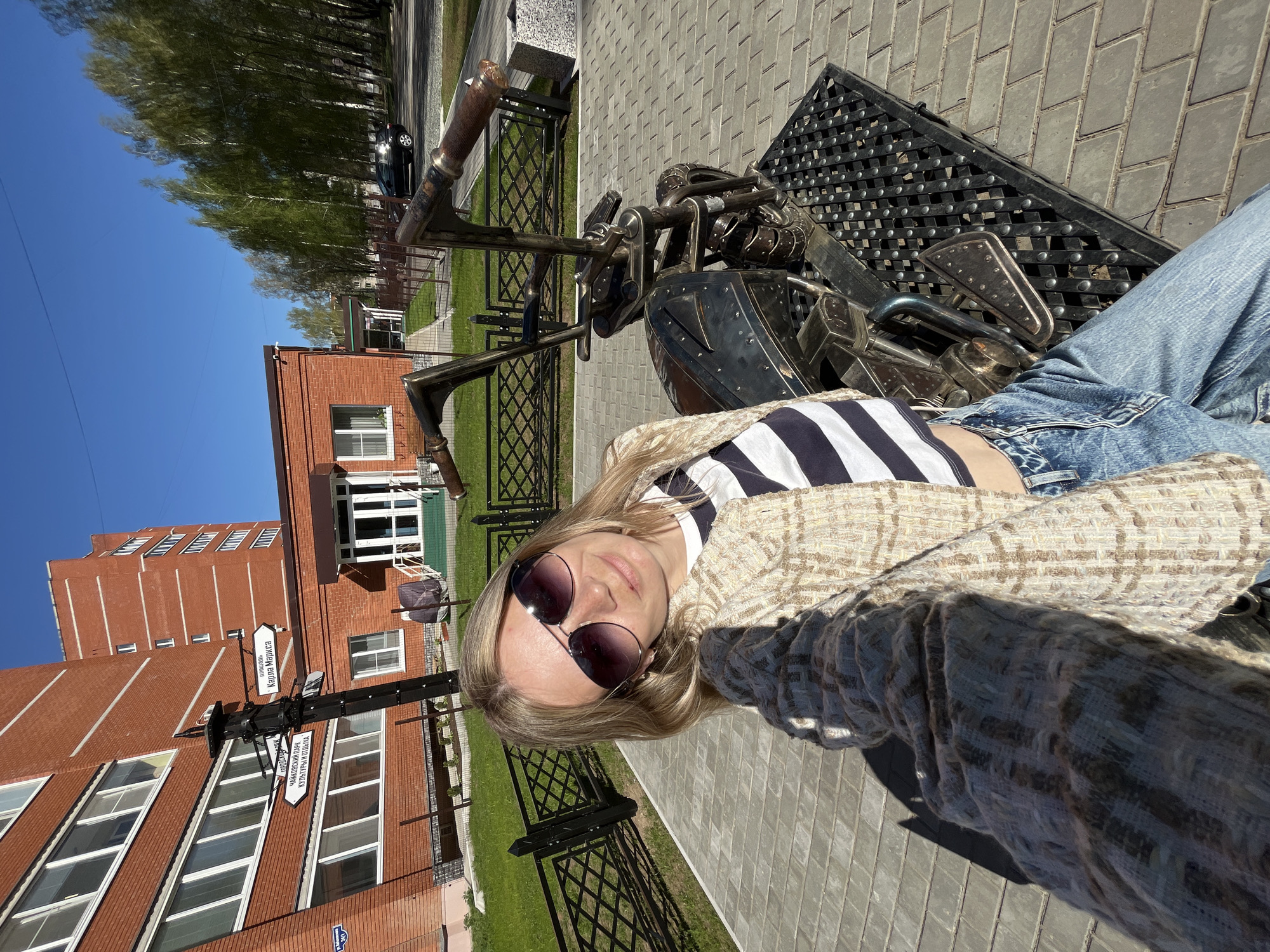 A selfie shows a woman with long blonde hair and sunglasses, lying on what appears to be a metal sculpture of a motorcycle. She is wearing a striped top, jeans, and a tan tweed jacket. The photo is taken outdoors on a brick-paved path, with a red brick building and greenery in the background. A signpost with street names in Russian is visible, including "Karl Marks Street" and signs to a local park. The location appears to be Chaikovsky, Russia.