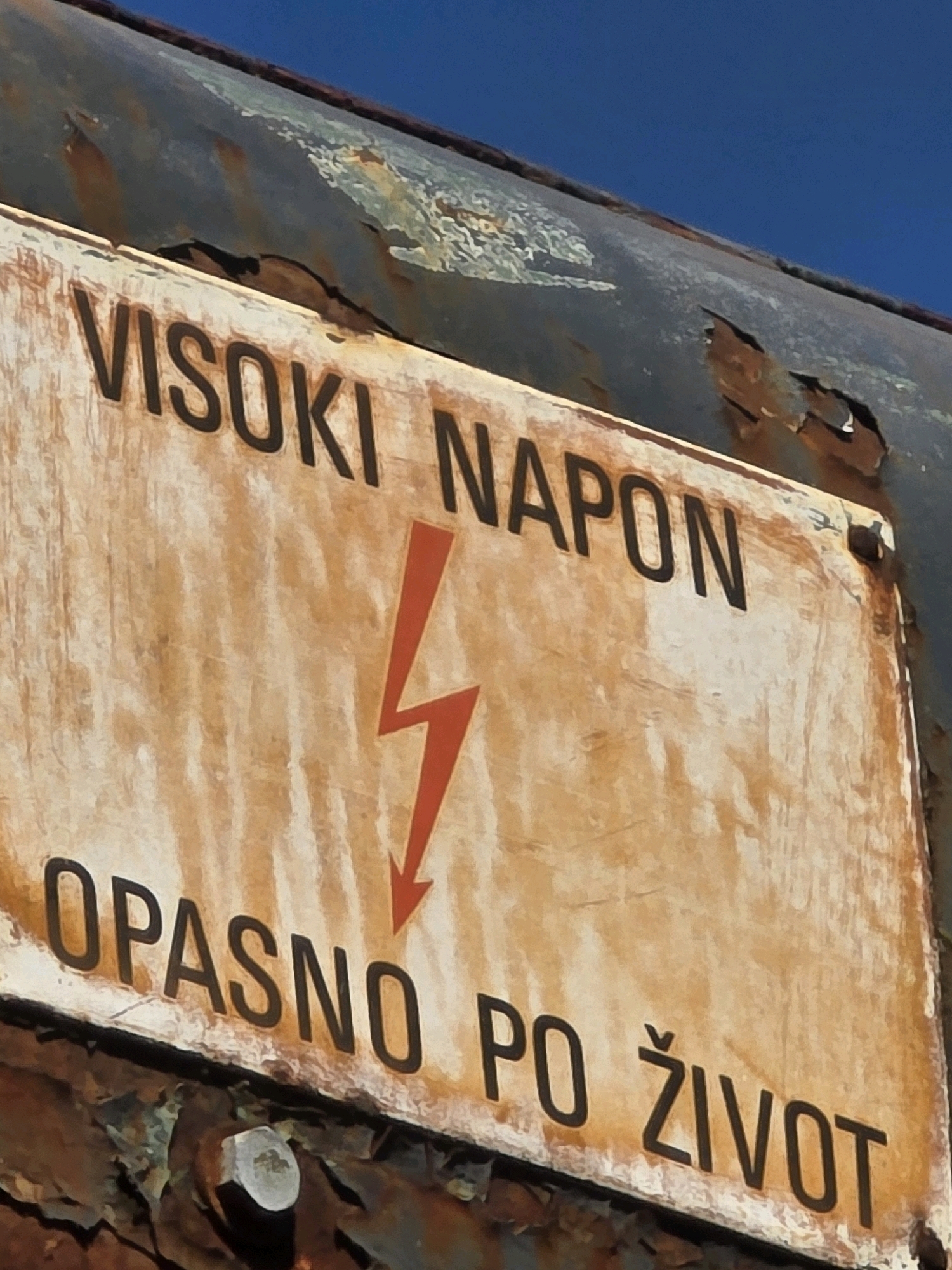 The image shows a weathered, rusted metal sign with black lettering and a red lightning bolt symbol. The text on the sign reads "VISOKI NAPON" above the lightning bolt and "OPASNO PO ŽIVOT" below it. This translates to "HIGH VOLTAGE" and "DANGEROUS TO LIFE". The sign is attached to a rusted, curved metal surface, likely part of industrial equipment or infrastructure. The background shows a clear blue sky. The sign is located on an outdoor industrial site. The overall condition of the sign and its mounting suggests exposure to the elements and a potentially hazardous environment.