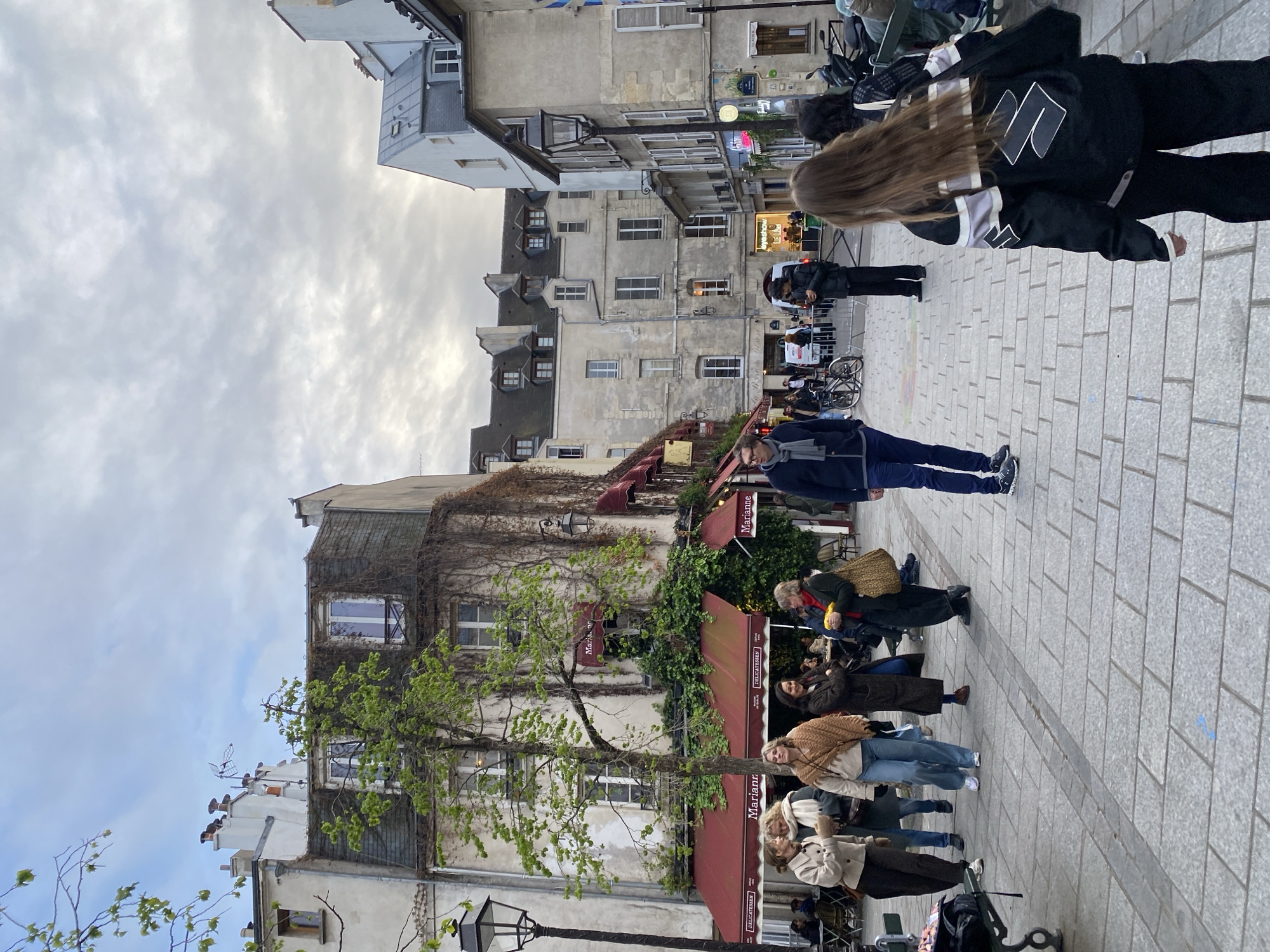 The image depicts a bustling pedestrian street scene on Rue des Hospitalières Saint-Gervais in the Le Marais district of Paris, France. The street is paved with light grey rectangular pavers.
In the immediate foreground, the back of a person with long brown hair, wearing a black jacket with white and grey stripes and "Suzuki" branding, and black pants, is visible as they walk away from the viewer. To their left, in the mid-ground, a man in a dark blue jacket, grey scarf, blue jeans, and dark sneakers stands, facing slightly right. Further left, a group of six women is walking towards the viewer and interacting. One woman wears a light brown jacket and dark pants, another has a light scarf and dark coat, a third is in a light top, blue jeans, and a large brown knitted shawl. A fourth woman in a dark long coat and dark curly hair looks towards the group, while a fifth, in a dark coat with red accents, holds a yellow object and a woven bag.
The left side of the street is dominated by a multi-story building featuring "Marianne Delicatessen" with red awnings and lush green ivy climbing its light stucco facade. A bare-branched tree with nascent green leaves stands in front of this building. A tall, ornate black street lamp post is prominent on the far left. The building exhibits classic Parisian architecture with numerous windows and a dark slate mansard roof with dormers.
Across the street, and receding into the background, other multi-story buildings with varying shades of light-colored facades and traditional windows line the thoroughfare. Another black street lamp post is visible on the right. In the background, further down the street, more people are visible, including one individual looking at a phone near a parked bicycle, and others seated at outdoor tables under awnings.
The sky is overcast with a blanket of grey and white clouds, providing diffuse natural light to the scene.