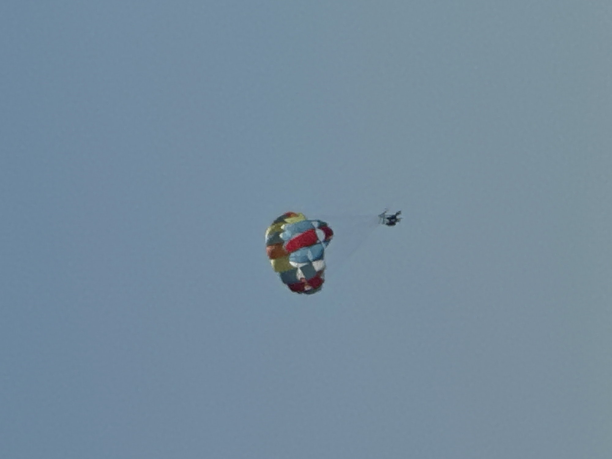 A brightly colored parasail, composed of red, blue, yellow, and green panels, is visible against a clear, pale blue sky. Two figures are suspended beneath the parasail by harnesses and ropes. The scene is outdoors with no discernible urban or natural landmarks, and thus cannot be located in Boreti, Montenegro.