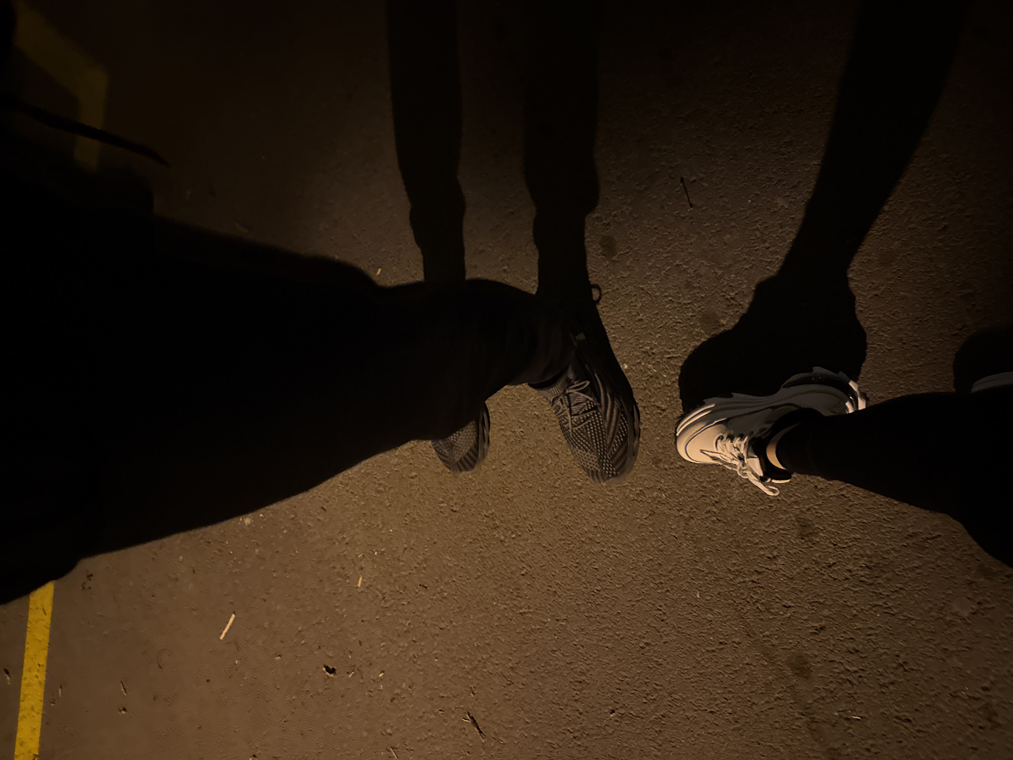 The image shows a low-angle perspective of three pairs of feet standing on a dark asphalt surface. The feet are wearing different types of shoes: one pair is wearing black knit running shoes, another pair is wearing white sneakers, and the third is indistinct due to darkness. The surface is illuminated enough to reveal the texture of the asphalt and shadows cast by the figures. A yellow line is visible in the lower left corner. The setting appears to be outdoors at night.