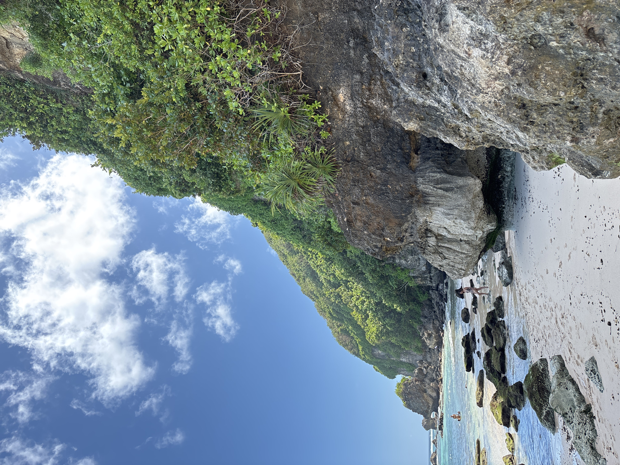 The image depicts a beach scene in Kuta Selatan, Indonesia. A rocky cliff covered in lush green vegetation dominates the upper portion of the frame. Below, a sandy beach stretches along the coastline, dotted with moss-covered rocks. The ocean water is clear and turquoise. In the middle ground, a person wearing a hat and swimsuit is walking along the shore. Further out in the water, another person is on a paddleboard. The sky is blue with scattered white clouds.