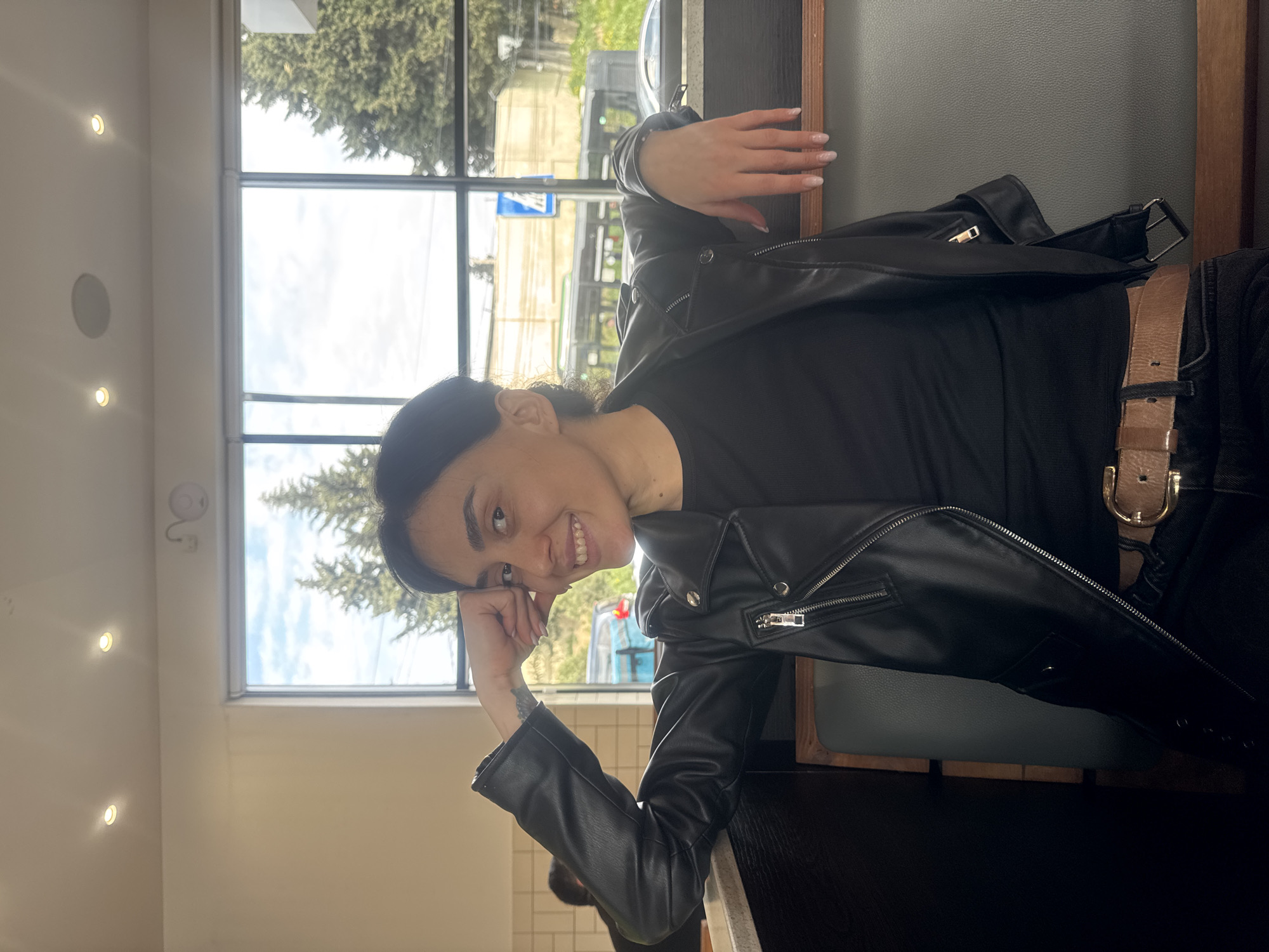 The image captures a young woman smiling warmly at the camera, seated within an indoor setting that appears to be a cafe or restaurant in Tbilisi, Georgia. She has dark hair pulled back and is dressed in a black leather-style jacket over a black top, paired with dark bottoms and a prominent brown belt featuring a gold buckle. Her right hand gently supports her chin, while her left arm rests casually on the dark tabletop or booth partition.
Behind her, large windows reveal an outdoor scene with lush green trees and a partly cloudy sky, indicating daytime, possibly late morning or early afternoon. A blue rectangular sign with indistinct white text or symbols is visible through the window, suggesting a street or public area outside. The interior features light-colored walls adorned with recessed spotlights and a light gray-green upholstered booth seat. The overall atmosphere conveyed is one of relaxation and cheerfulness.