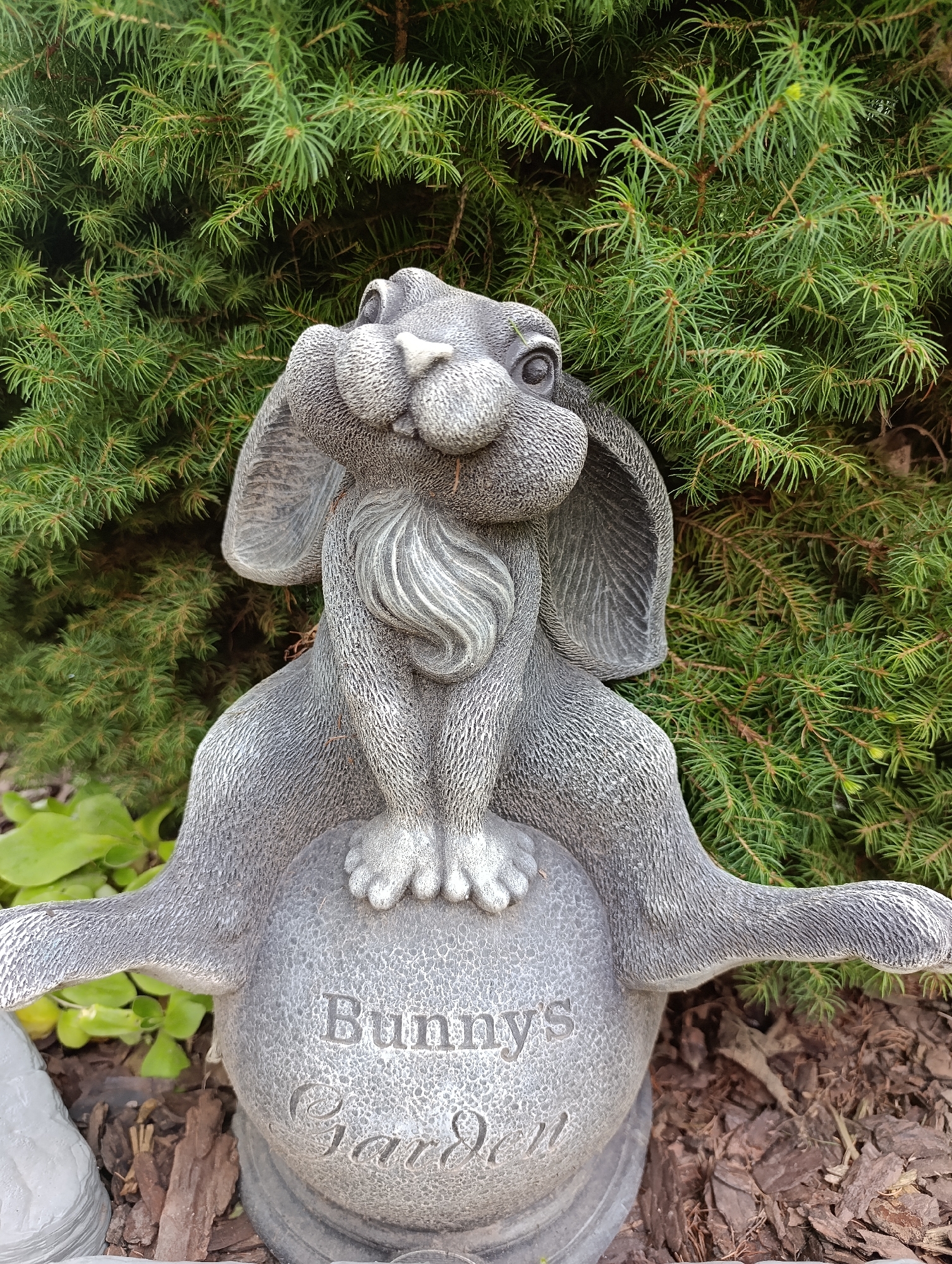 A stone statue of a bunny is positioned outdoors, nestled against a backdrop of dense green evergreen branches. The bunny statue is seated atop a spherical base inscribed with the words "Bunny's Garden." The statue is detailed, with textured fur and floppy ears. The ground around the base is covered in mulch. The image does not provide any information that would allow for the determination of its location in Kommunarka, Russia.