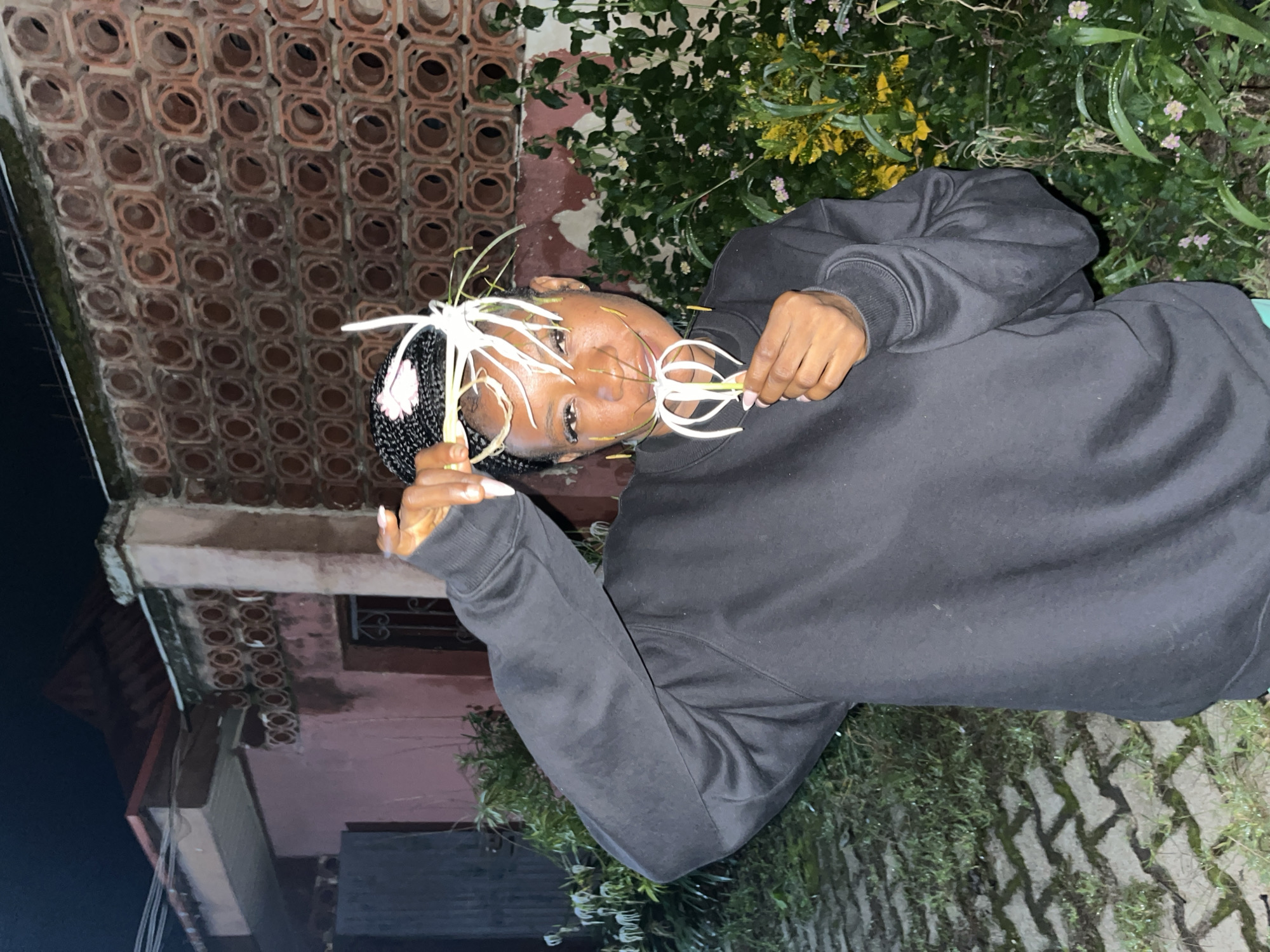 A young woman with dark skin and braided hair is standing in front of a building with a patterned brick wall. She is wearing a black sweatshirt. She is holding white flowers with long tendrils up to her face, and she appears to be smiling. The ground is paved with interlocking bricks and there is greenery in the background. The image is dimly lit, suggesting it was taken at night. The architecture and vegetation are consistent with the climate and style found in Jos, Nigeria.