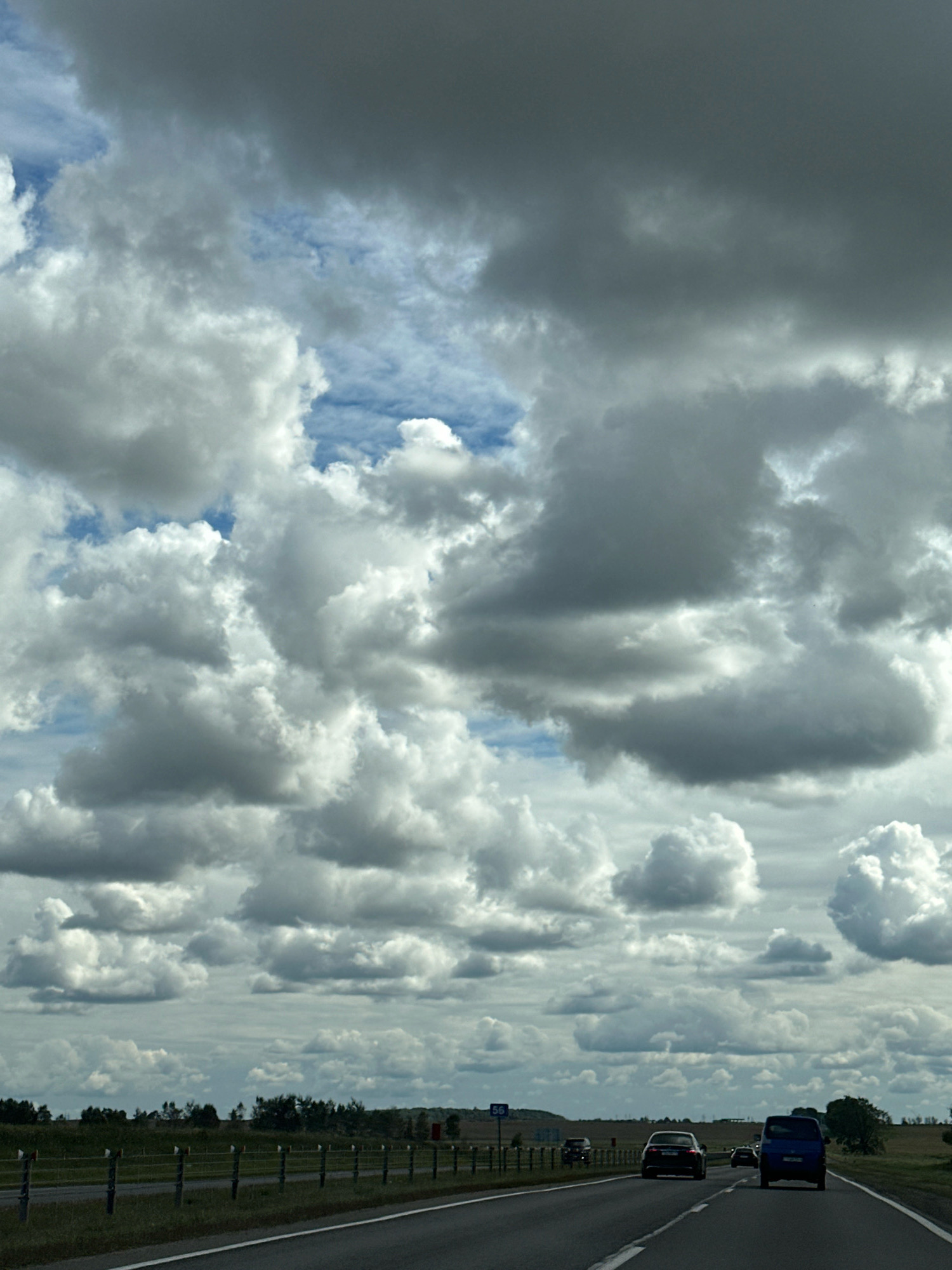 The image depicts a view from a moving vehicle on a highway, likely within the Блонскі сельскі Савет region of Belarus. The sky is dominated by a dramatic and expansive cloudscape, with large, fluffy white cumulus clouds interspersed with darker, more ominous storm clouds. Patches of blue sky are visible between the clouds.
The highway is asphalt with white lane markings. In the foreground, the road curves slightly to the right. Several vehicles are traveling on the highway. A dark-colored sedan is in the right lane, ahead of the viewer. A blue van is in the left lane, further ahead. Other vehicles are visible in the distance.
To the left of the highway, a metal fence with vertical posts and horizontal wires runs along the roadside, separating the road from a grassy embankment. Beyond the embankment, a flat, rural landscape unfolds, characterized by green fields and scattered trees. In the far distance, a line of trees forms a treeline against the horizon. A red rectangular structure, possibly a building or a sign, is visible in the far right background. A blue road sign with the number "56" is also visible on the left side of the road. The overall impression is of a journey through a rural Belarusian landscape under a dynamic sky.