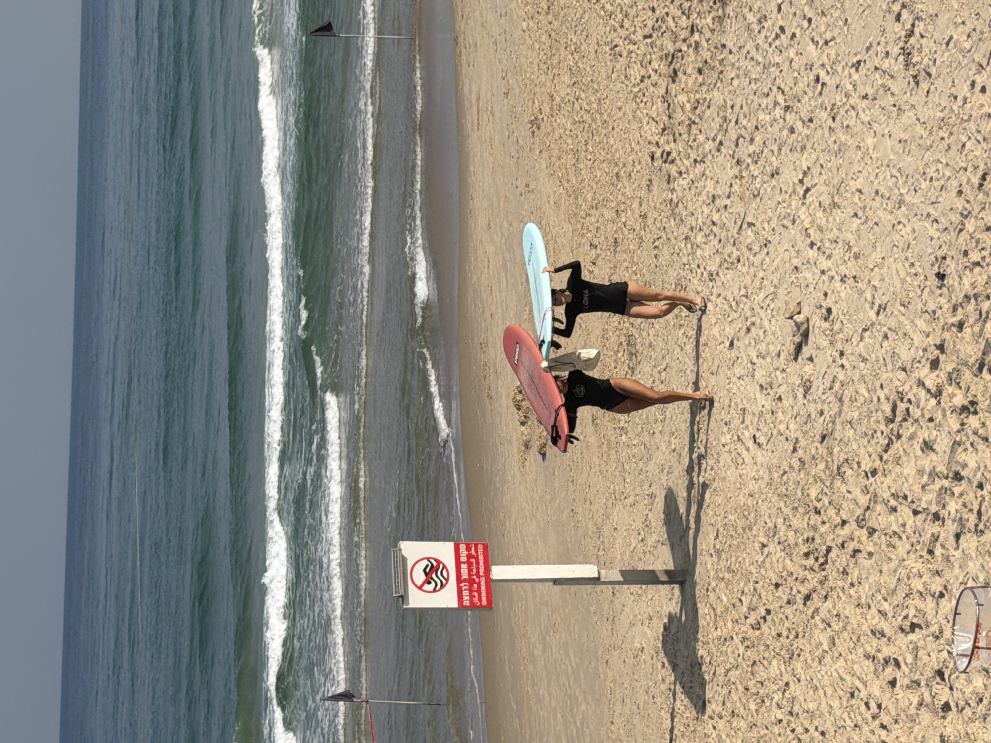 The image depicts a sunny day at a beach in Tel Aviv, Israel. In the foreground, two young women are walking on the sand, each carrying a surfboard above their heads. The woman on the left is wearing a black swimsuit and carrying a red surfboard. The woman on the right is also wearing a black swimsuit and carrying a light blue surfboard. Behind them, a red and white sign is posted on a pole, which reads "SWIMMING PROHIBITED" in English and Hebrew, indicating this area of the beach is not safe for swimming. Further back, the turquoise waves of the Mediterranean Sea are visible, with white foam cresting on the water's surface. The sky is a clear blue, suggesting good weather conditions. The beach is made of light-colored sand, with some footprints and debris scattered across its surface. A black flag on a pole is planted in the sand near the water's edge, possibly indicating a flag warning or a lifeguard station. The overall scene suggests a recreational setting, despite the prohibition of swimming.
