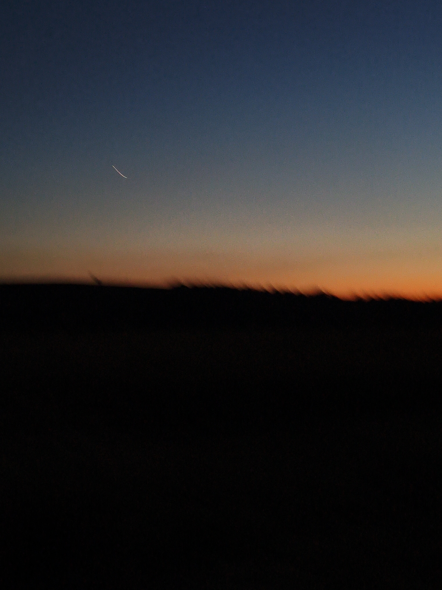 This is a photograph of a sky at dusk. The sky is a gradient of dark blue at the top, transitioning to orange and then yellow closer to the horizon. A faint, curved line, likely a contrail from an airplane, is visible in the upper left quadrant of the image. Below the sky, a dark, silhouetted landscape stretches across the bottom half of the frame. The horizon line appears jagged and blurred, suggesting movement or a stylized rendering of the land. The overall mood is serene and peaceful, characteristic of the transition from day to night. The time of day is clearly dusk or twilight. No people, objects, visible text, or specific location cues are discernible in the image.