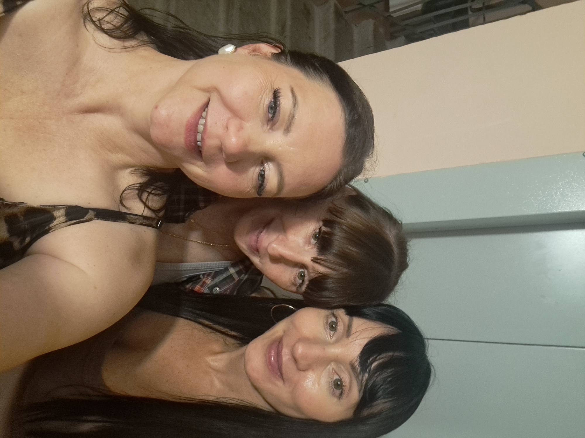 Three individuals are posing for a selfie indoors, with a pale pink wall on the left and a grey vertical surface on the right. The person in the foreground on the left is a woman with dark brown hair and fair skin, wearing a leopard print top and a pearl earring. She is smiling and looking towards the camera. Behind her and to the right are two other women. The woman in the middle has brown hair with bangs and is wearing a plaid shirt and a grey tank top. The woman on the far right has long black hair and is wearing a brown tank top. She is also looking towards the camera. The background appears to be a hallway or interior space.