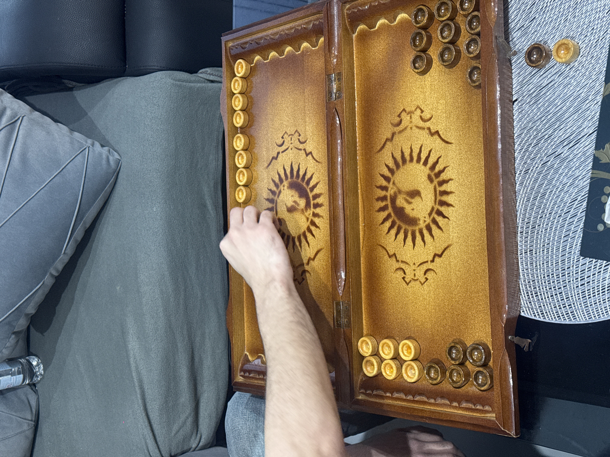 An indoor scene shows a person's arm and hand reaching onto an open backgammon board, suggesting a game in progress or being set up. The setting appears to be a comfortable living area, possibly in Yerevan, Armenia, given backgammon's popularity in the region.
The primary subject is a highly decorative wooden backgammon (nard) board, open and lying flat. The playing surfaces are a rich, golden-brown hue with darker brown, intricately carved sun motifs and other stylized patterns in the center of each half. Light-colored (cream/yellow) and dark-colored (brown) checkers are arranged on the board. The left side of the board shows several light checkers stacked neatly along the outer edge, while the person's hand is positioned near the playing field with more light checkers. The right side of the board has both light and dark checkers, some stacked, along its outer edge and playing field.
The board itself is made of wood, likely varnished or lacquered, with decorative scalloped edges and a central hinge. To the left of the board, a gray, soft throw pillow with pleated detailing rests on what appears to be a dark gray or black sofa or chair. A small, clear plastic water bottle is partially visible near the pillow. To the right of the board, part of a textured white or silver mat or table covering is visible, on which a couple of checkers are resting, adjacent to a dark surface, possibly a table. No specific text is clearly visible on any objects in the image. The time of day is not discernible, but the scene is well-lit, likely by indoor artificial light.