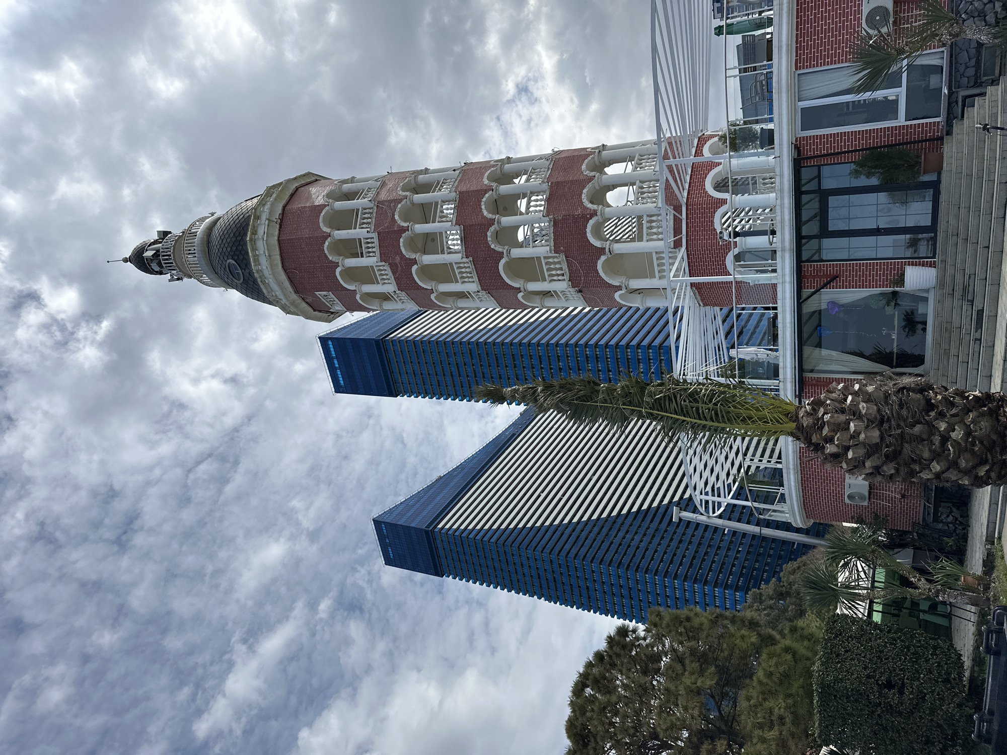 This image shows two modern, blue-glass skyscrapers with slanted tops and the Alphabetic Tower in Batumi, Georgia. The tower, featuring red-brick and white-column architectural elements, is accompanied by an attached building, palm trees and other vegetation. The sky is cloudy.