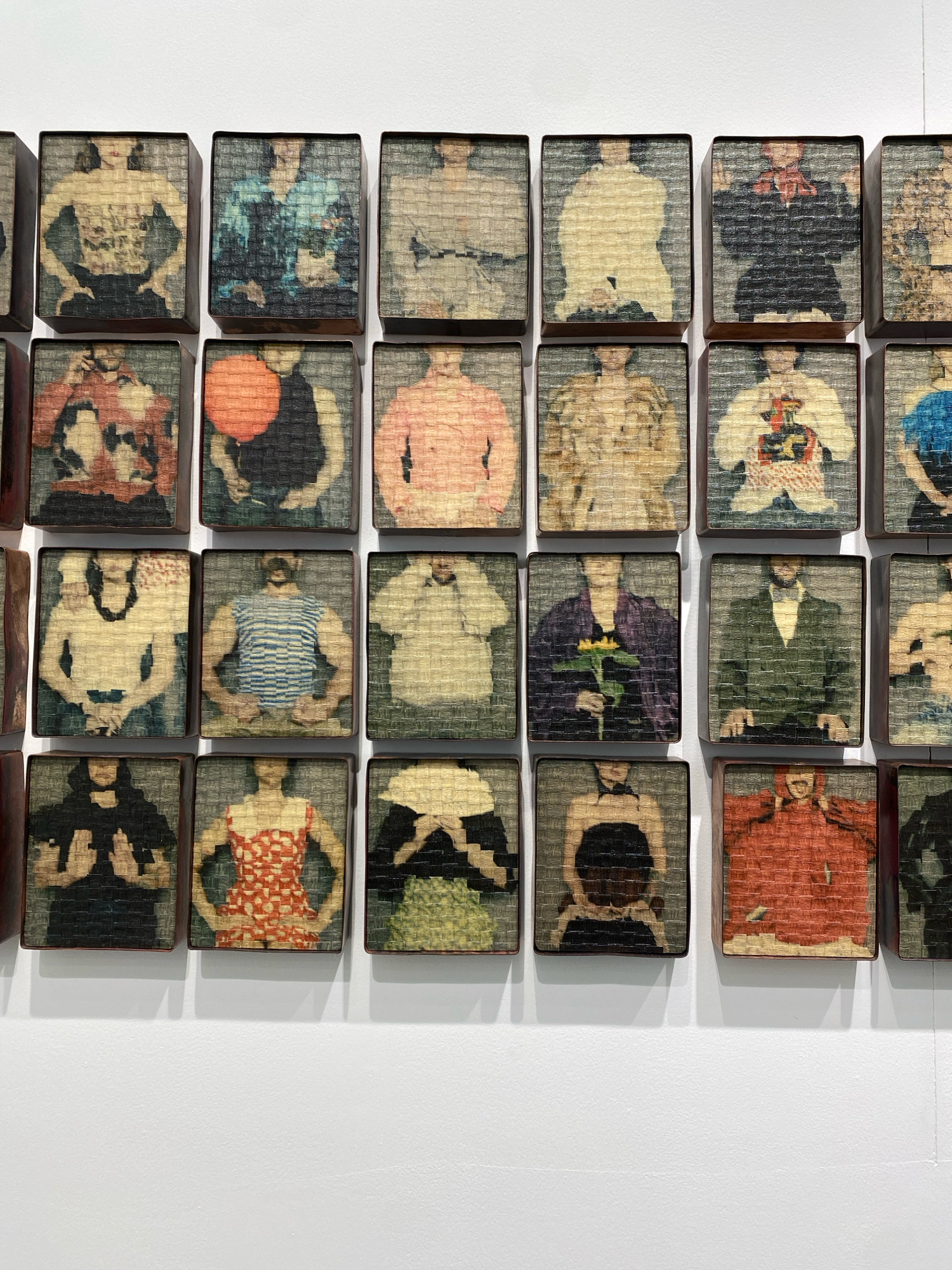 The image displays a grid of small, square artworks mounted on a white wall. Each artwork appears to be a framed photograph or print of a person, depicted from the chest up. The style of the artworks is consistent, with a textured, almost woven appearance. They are arranged in five rows and six columns, with some being partially visible at the edges. The artworks are predominantly in muted tones, with some featuring brighter colors like red, blue, and yellow. The lighting suggests an indoor setting, likely an art gallery or exhibition space. The background is a plain white wall, and shadows are cast below the artworks, indicating a light source from above. There is no identifiable text or signage within the frame. The environment appears clean and well-lit, typical of a gallery.