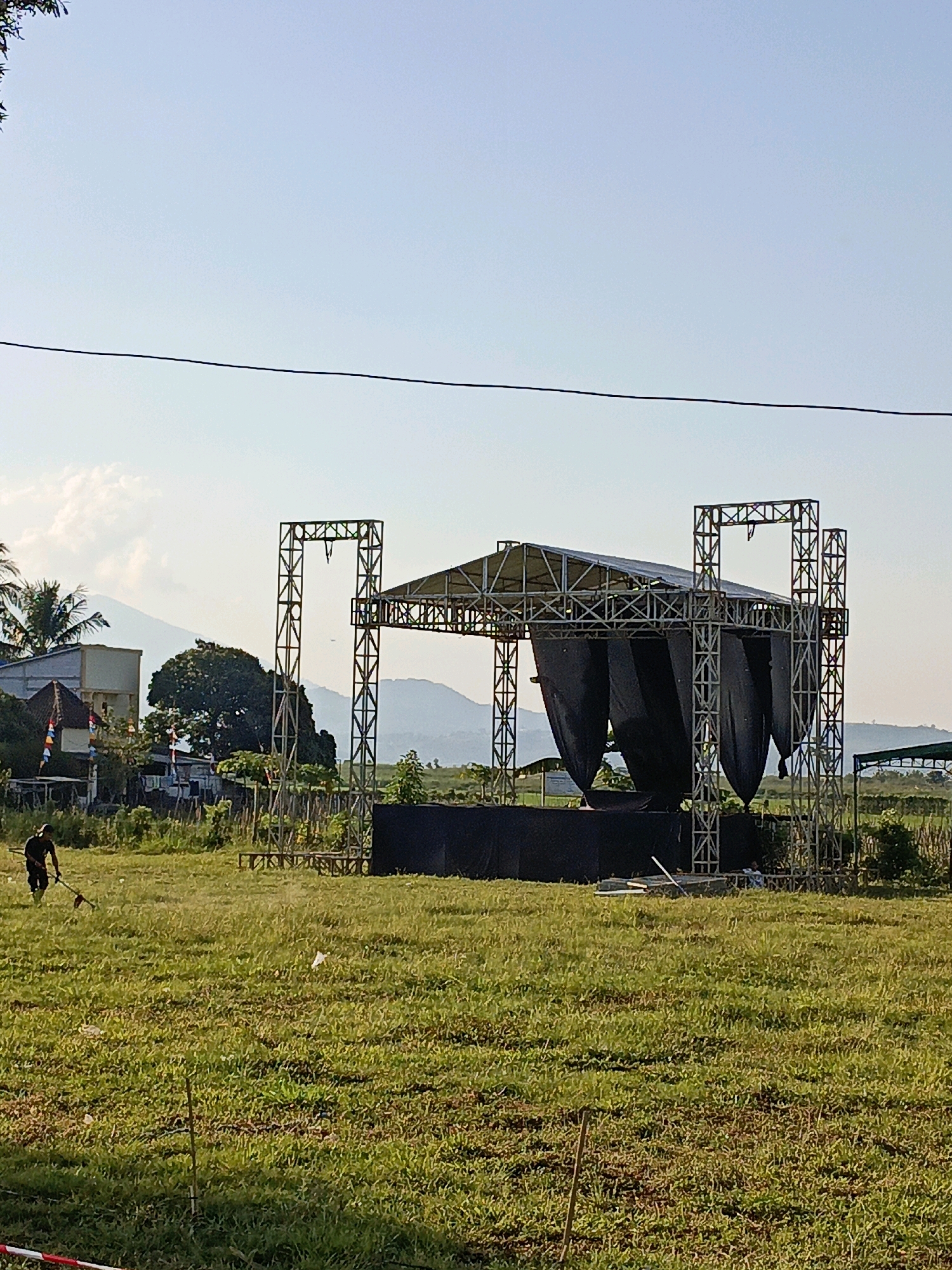 The image depicts an outdoor scene in Rowosari, Indonesia, featuring a temporary stage structure erected in a grassy field. The stage is constructed from metal trusses and covered with black fabric, suggesting preparation for an event. In the foreground, a person is operating a grass trimmer, indicating ongoing site maintenance. The background reveals a landscape with trees, buildings, and distant hills under a clear sky. The overall impression is one of outdoor event preparation or setup.