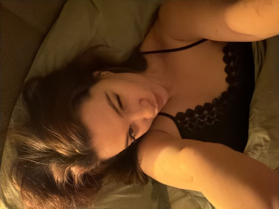 A woman with brown hair rests against a green pillow. She wears a black tank top with lace detailing along the neckline. She lies on her side, her face angled towards the camera, eyelids closed. Soft lighting illuminates her skin and hair. Her arm extends towards the camera, creating a sense of intimacy and immediacy. The overall image is relaxed and cozy, suggestive of a moment of rest or sleep.